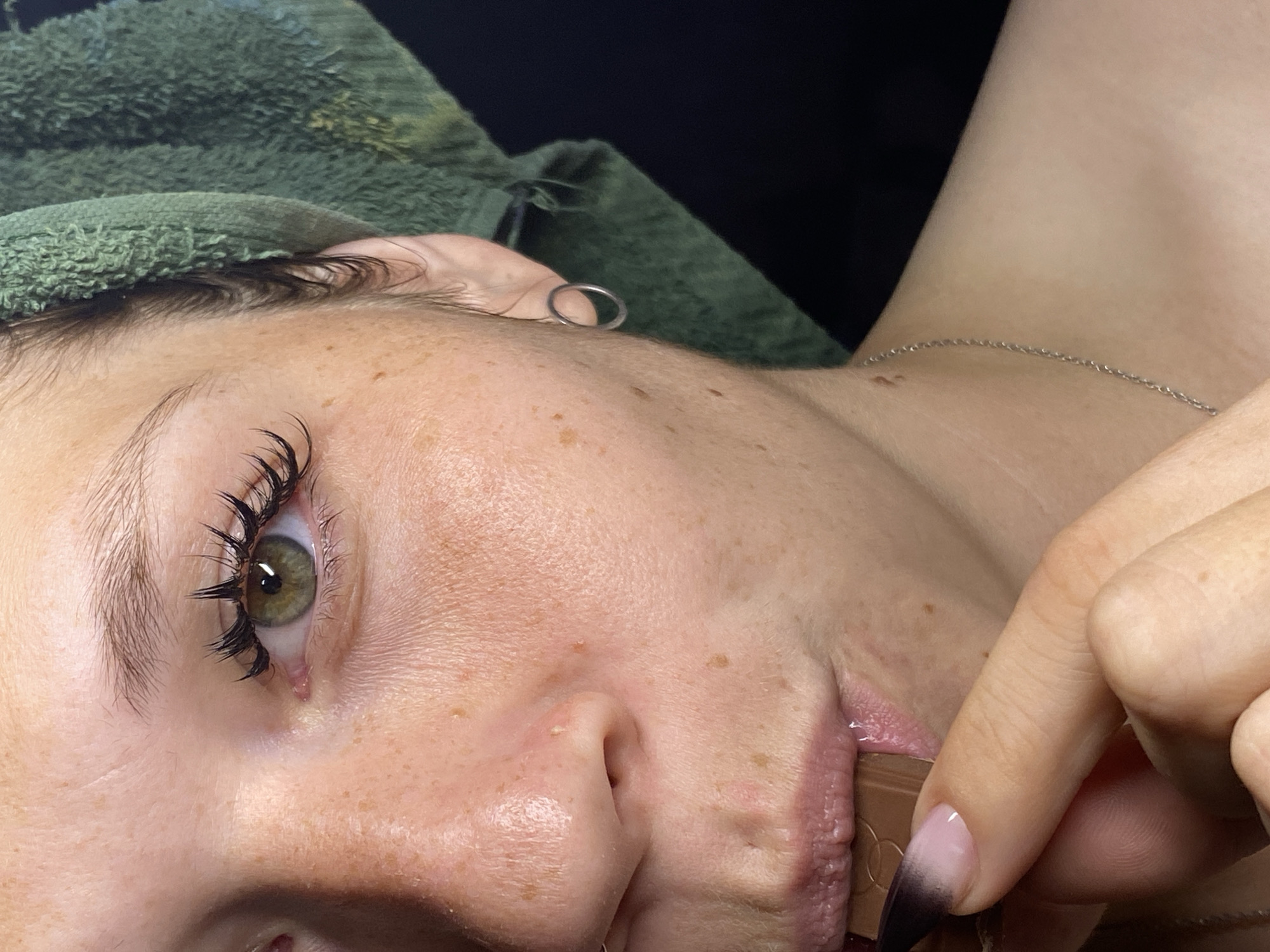 This is a close-up, upside-down, over-the-shoulder shot of a person's face, likely in a beauty salon setting in Chaikovsky, Russia. The subject has fair skin with freckles, green eyes with dramatic false eyelashes, and light eyebrows. A green towel is draped over their head and shoulders. A thin silver necklace is visible around their neck. The subject's right hand is holding a piece of chocolate up to their mouth. The nails on this hand are long and have a gradient of pink to black. The lighting suggests an indoor environment, possibly with artificial light, and no specific time of day or weather is discernible. The emotion conveyed is subtle, possibly one of enjoyment or anticipation of eating the chocolate. There is no visible text in the image.
