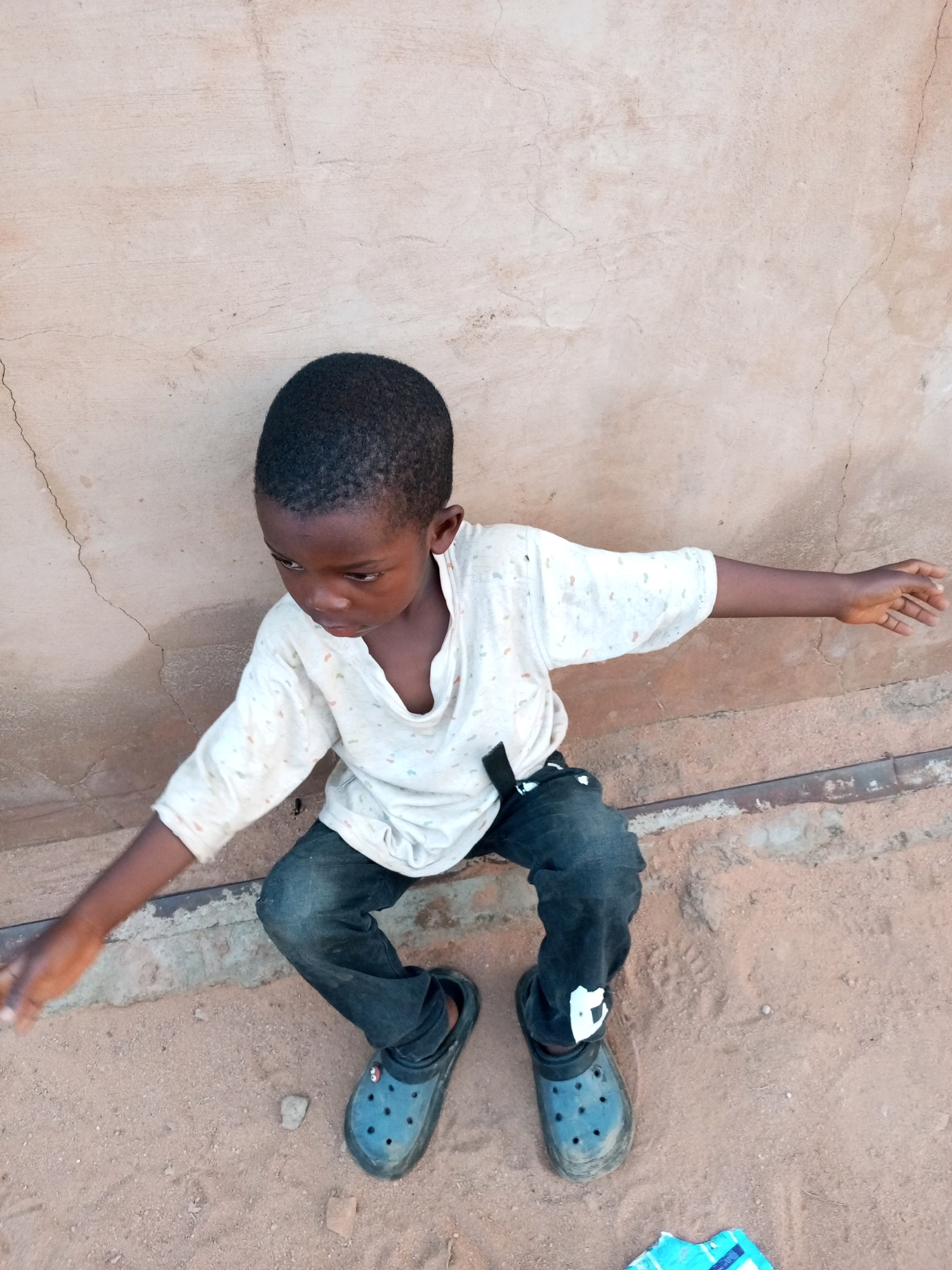 A young Black boy is sitting on sandy ground leaning against a light brown, cracked, cement wall. He is wearing a light-colored, short-sleeved shirt with a subtle all-over pattern of small colorful dots, and dark denim jeans. He has on blue Croc-style shoes. His arms are extended to his sides. He is looking downwards. The background wall shows signs of wear and tear with visible cracks. The ground is composed of fine sand with some small stones and debris. A thin strip of concrete or metal runs horizontally across the lower part of the image behind the boy.