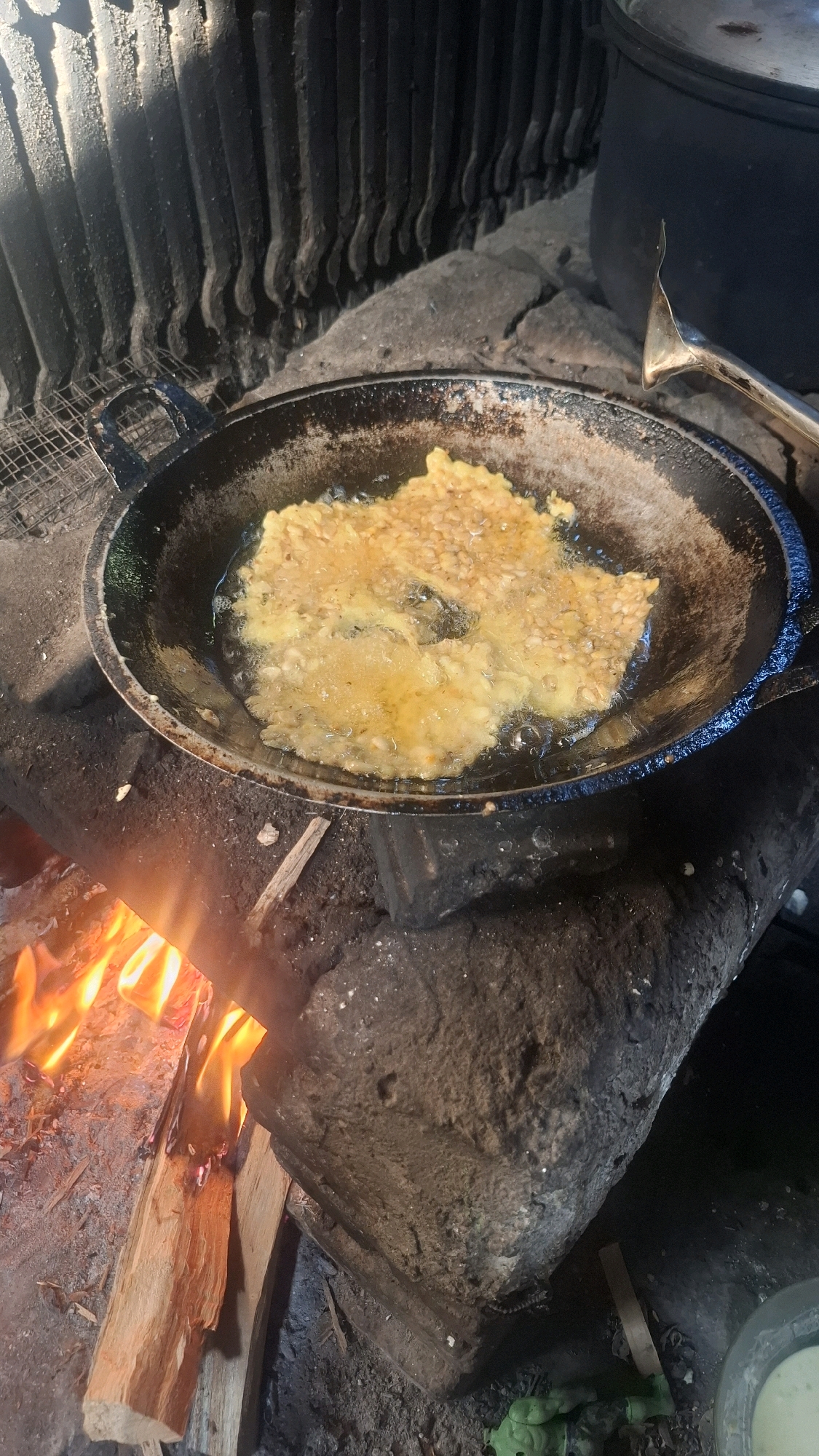 In the village of Semanding, Indonesia, a close-up shot captures the process of frying tempeh over an open wood fire. The tempeh, coated in batter, sizzles in a dark, well-worn wok resting on rough, dark stones. Bright orange flames lick the bottom of the wok, indicating intense heat. In the background, a traditional cooking setup is visible, with dark, textured walls that appear to be made of clay or soot-covered brick, and a large dark pot to the right. A metal tong rests near the wok, suggesting active cooking. The overall scene evokes a rustic, traditional method of food preparation.