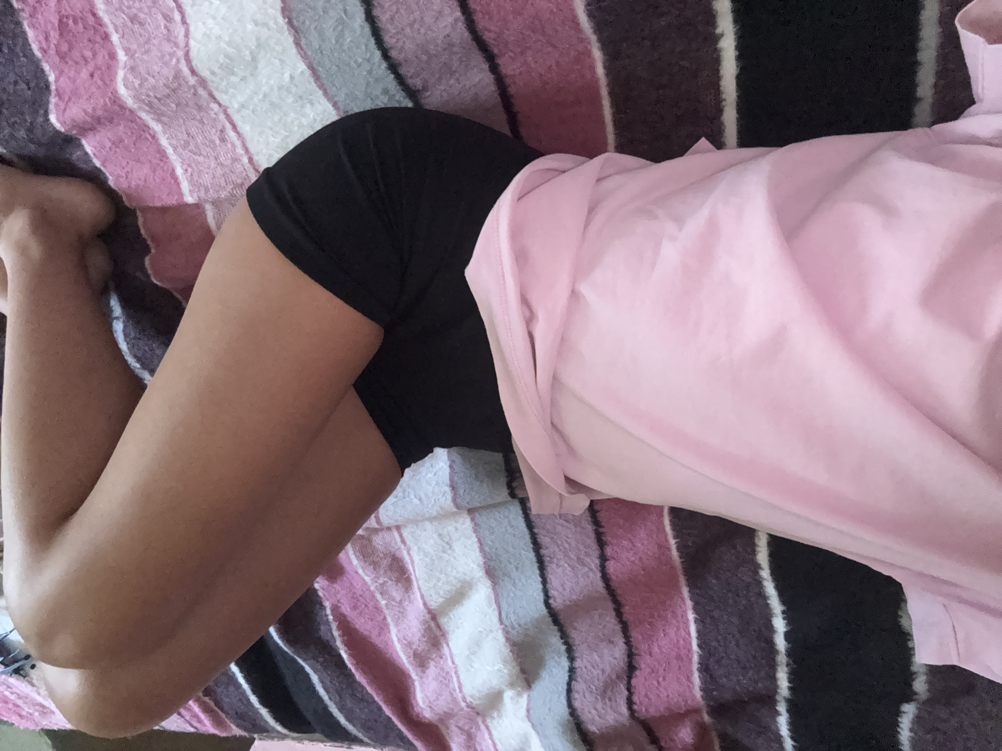 A person is lying on a bed with striped bedding. The person is wearing black shorts and a pink shirt that is draped over their body. The bedding has stripes of pink, white, and dark gray. The person's legs are bent, and their feet are visible on the left side of the image. The lighting suggests it is daytime. The scene is indoors, likely a bedroom.