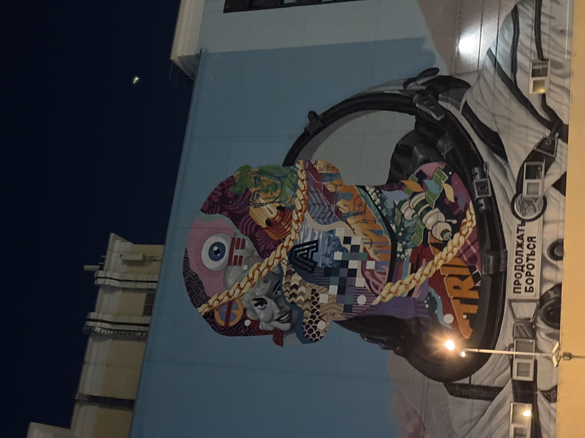 This image shows a large mural on the side of a building. The mural depicts an astronaut's helmet filled with various artistic motifs, including a stylized eye, portraits of famous figures like Marilyn Monroe, geometric patterns, and abstract shapes. A gold chain wraps around the "head" portion. The astronaut's suit is visible around the helmet. The text in Russian, "ПРОДОЛЖАТЬ БОРОТЬСЯ," (PRODOLZHAT' BOROT'SYA) meaning "Continue to Fight" or "Keep Fighting", is seen on the astronaut's suit. The mural is illuminated by streetlights. The photo was taken in Moscow, Russia.