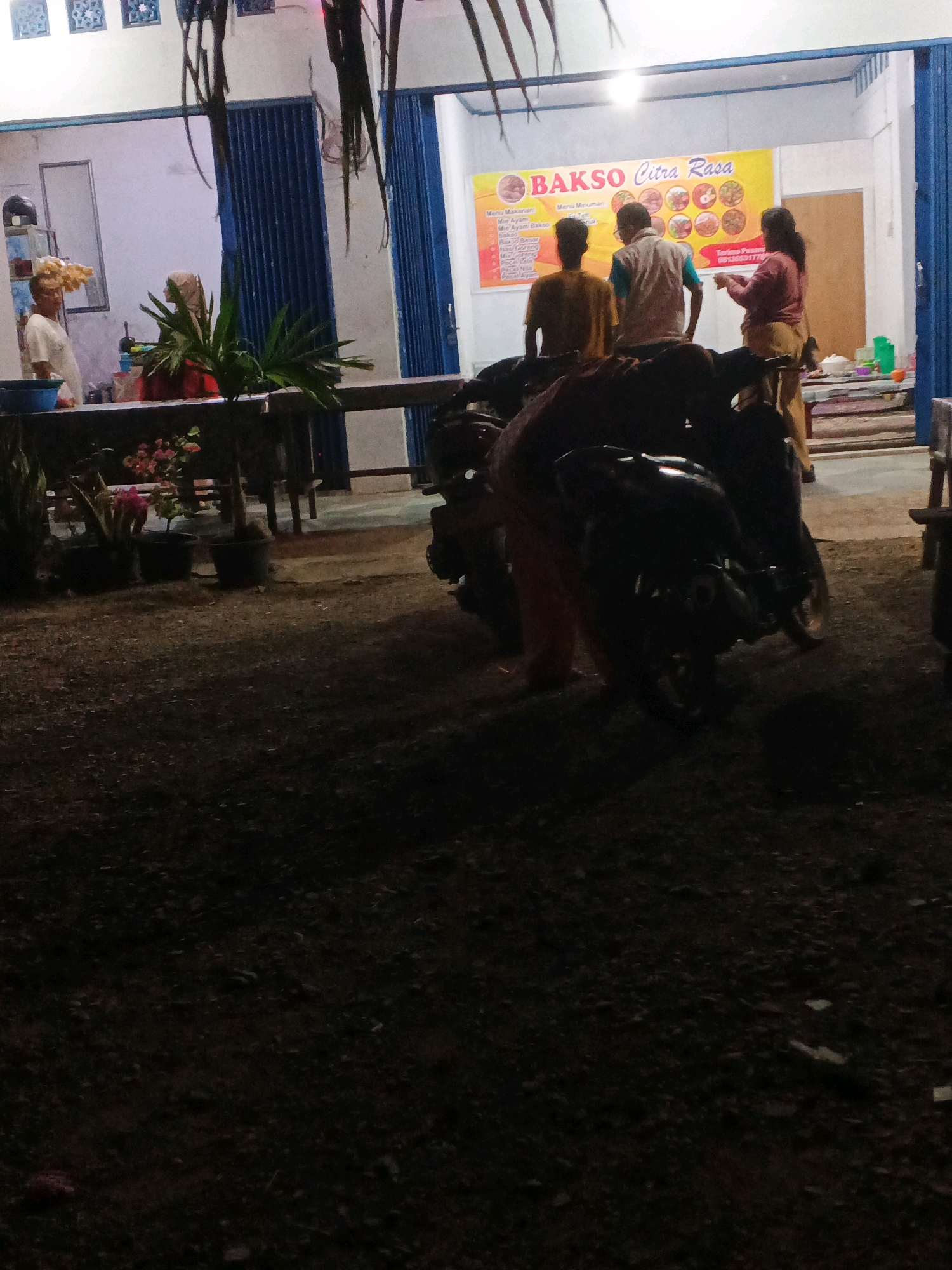 The image depicts an outdoor scene at night, likely in Brangan Mulya, Indonesia, featuring a food stall and its surroundings. In the background, a brightly lit food stall with a yellow banner that reads "BAKSO Citra Rasa" is visible. The banner also includes images of food and text in Indonesian, possibly a menu or advertisement. Three individuals are near the stall; one, wearing a yellow shirt, stands with their back to the camera. Another, in a white shirt and vest, faces away from the camera, while a third, in a pinkish-brown top and tan pants, faces the stall.
In the foreground, a black motorcycle is parked, partially obscuring the lower portion of the stall. The ground is covered with gravel and dirt. Several potted plants with green foliage and some with pink flowers are arranged in front of the stall. The overall lighting suggests it is evening or night, with the stall being the primary source of illumination in its immediate vicinity. The background also shows a blue rolling door and part of a white building.