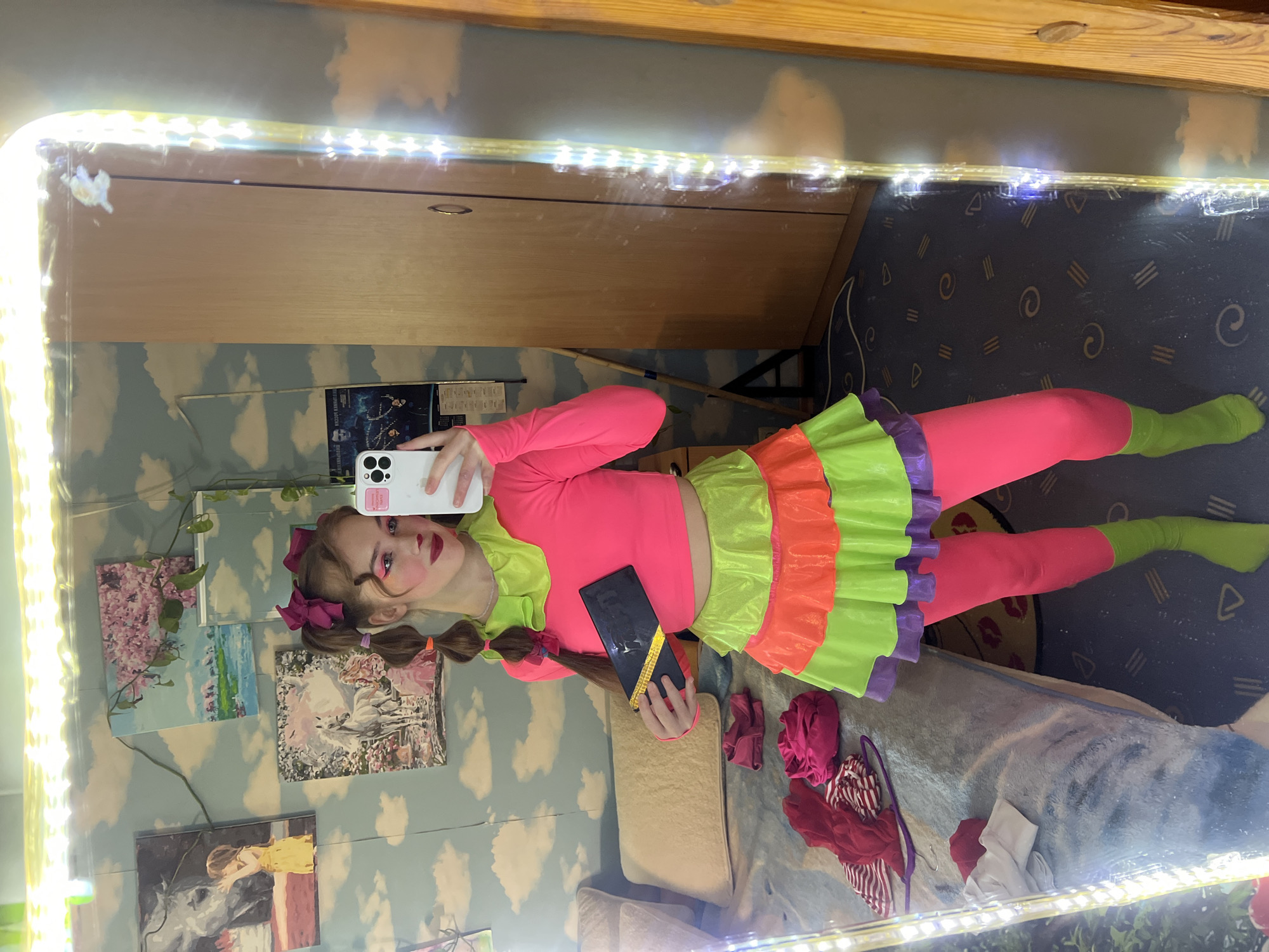 A young person in a vibrant clown-like outfit (pink top and tights, green socks, ruffled skirt of green, orange, and purple) is taking a selfie in a mirror framed with bright lights. The person is holding a smartphone in one hand and a dark object (possibly a makeup palette) in the other. They have pigtails with magenta bows, and their makeup is bold with bright pink eyeshadow and red lipstick. The room in the reflection has wallpaper patterned with clouds, and several framed pictures are visible on the wall. There is a wooden wardrobe in the upper background and patterned carpeting on the floor. Additional clothing items are scattered on the bedding below the person. The location is implied as Chaikovsky, Russia, based on the prompt.