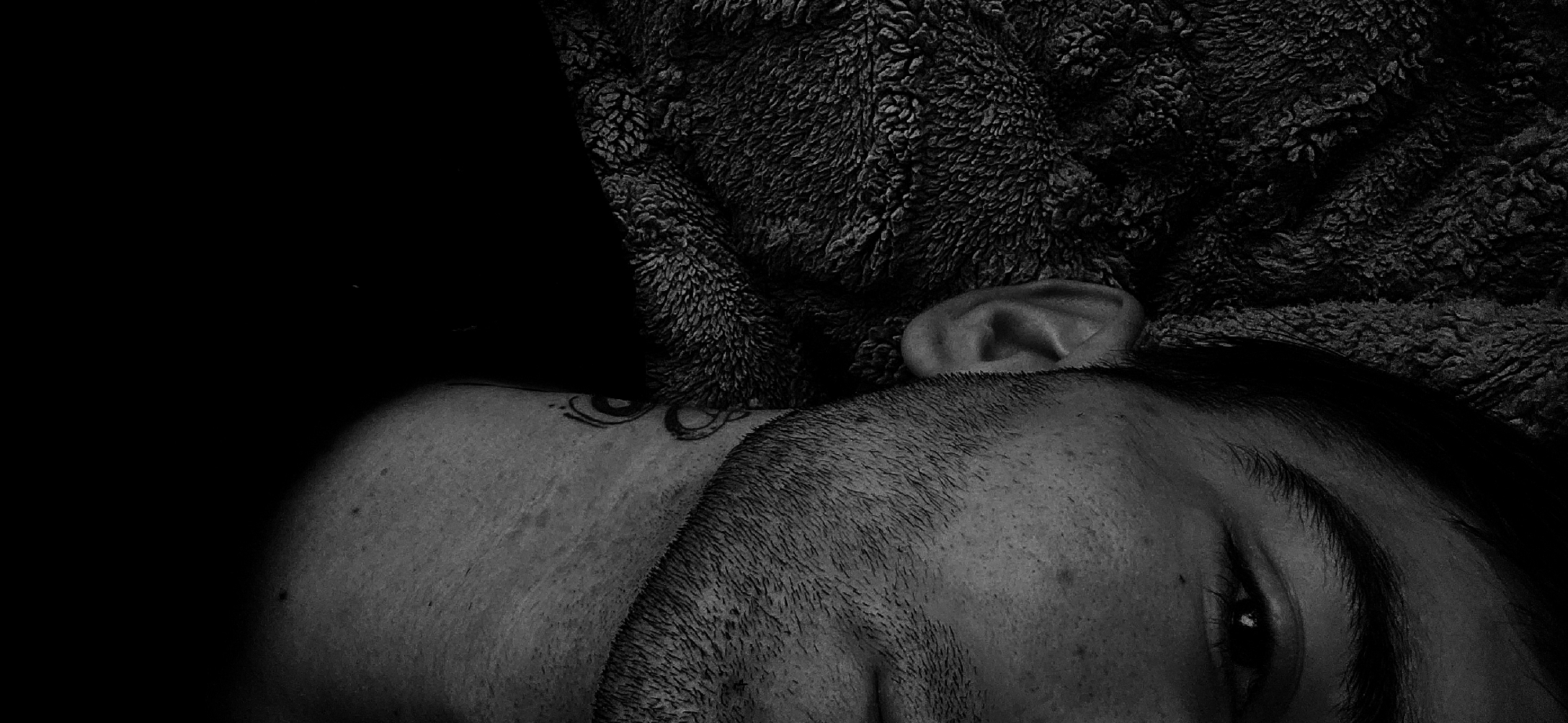 A black and white close-up shot captures a person lying down, possibly resting or sleeping. The image is tightly framed, focusing on the upper part of the person's head and shoulder. A prominent feature is the textured, fluffy fabric, likely a blanket or a hooded garment, that envelops parts of the head and neck, providing a strong textural contrast against the skin.
The person's ear is clearly visible, peeking out from beneath the fabric. Below the ear, stubble or very short hair is noticeable on the side of the head or face, suggesting a close shave or facial hair. A small, intricate tattoo with swirling, abstract elements is visible on the person's neck or upper shoulder, partially obscured by the textured fabric.
A portion of the person's eye is visible towards the lower right of the frame, appearing relaxed or partially closed. The lighting is dramatic, creating deep shadows and strong highlights that emphasize the textures of the fabric, skin, and hair. The background is predominantly dark, enhancing the focus on the subject. There are no direct visual cues for the time of day or specific weather conditions, but the presence of the blanket suggests an indoor setting and a mood of comfort or introspection. No visible text is present in the image.