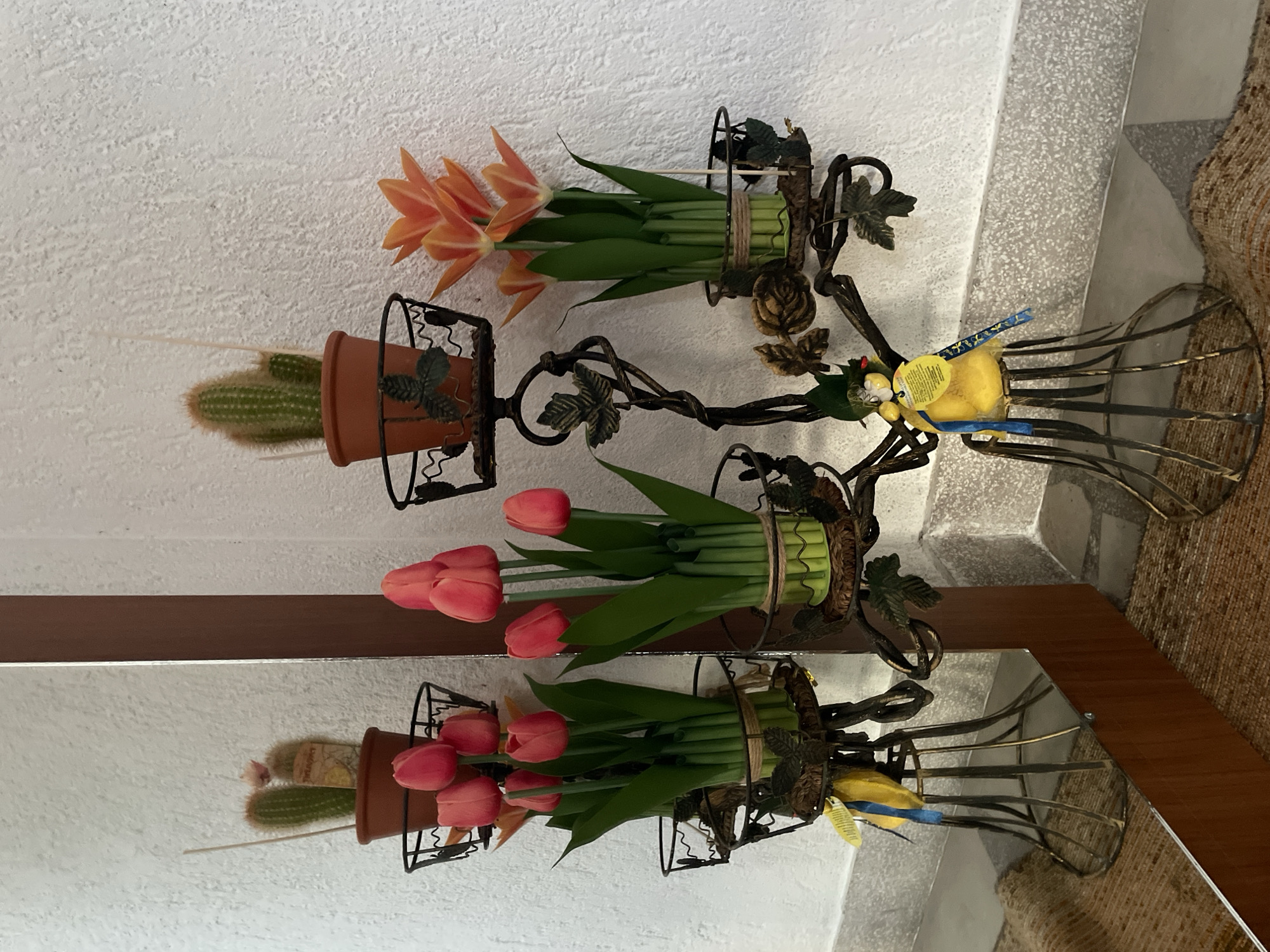 A multi-tiered, dark metal plant stand holds various potted plants and decorative elements. On the top tier, to the left, is a small potted cactus with a pink bloom. To the right on the top tier is another potted cactus. The middle tiers of the stand hold bouquets of red tulips in green holders, wrapped with twine. To the right of the red tulips, orange tulips are displayed in a similar green holder. The lower section of the stand features a decorative metal bird figurine adorned with blue and yellow ribbons. The plant stand is positioned against a textured white wall. To the left of the plant stand, a large mirror reflects the scene, showing another red tulip bouquet and the top of the cactus. The foreground is occupied by a textured brown rug and a light-colored tiled baseboard. The image appears to be taken indoors, possibly in a home or a public space in Zlatograd, Bulgaria, given the presence of the blue and yellow ribbons which may symbolize Ukrainian support.
