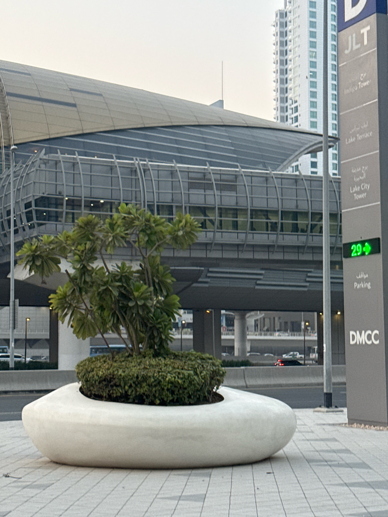 The image captures a street-level view of the DMCC metro station in Dubai, United Arab Emirates. In the foreground, a large, white, oval-shaped planter holds a small tree with lush green foliage, surrounded by dense, low-lying green bushes. The planter is situated on a tiled pedestrian walkway. To the right of the planter, a tall, grey directional sign stands, featuring Arabic and English text indicating "JLT," "Indigo Tower," "Lake Terrace," and "Lake City Tower." Below these destinations, the sign displays "Parking" in both languages, with a green digital display showing "29" and a right-pointing arrow, likely indicating parking availability or direction. Further down the sign, "DMCC" is prominently displayed. In the background, the elevated structure of the DMCC metro station, with its distinctive curved, silver roof and glass-paneled walkway, is visible. Beyond the station, a modern, white high-rise building with many windows stands tall against a hazy sky. A road with cars is visible in the middle ground.