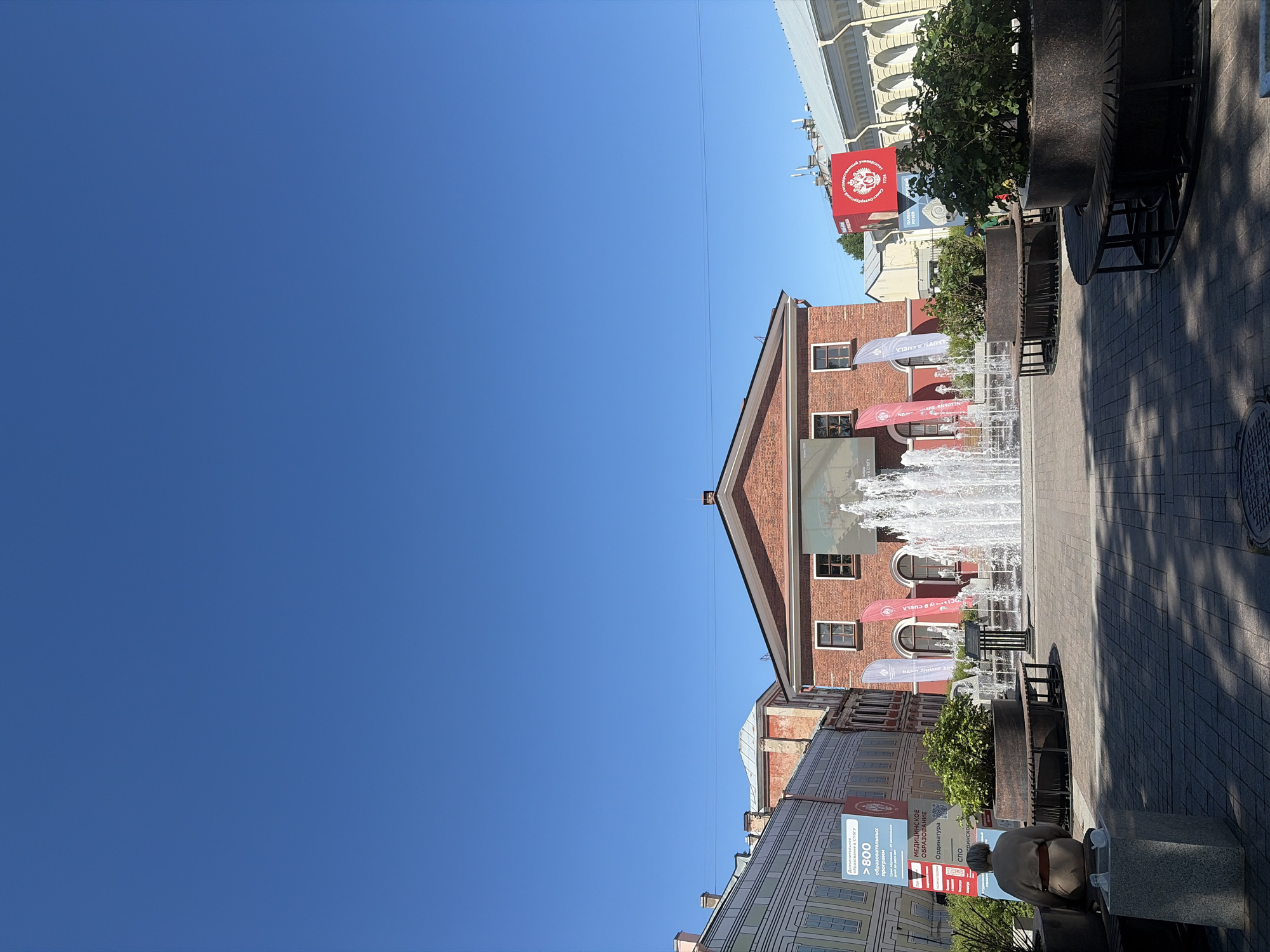 The image displays Muzeynaya Ploshchad (Museum Square) in Saint Petersburg, Russia, on a clear, sunny day. The primary focus is a pedestrian square fronting the red brick exhibition building of the "Russia - My History" Historical Park, located at Konyushennaya Square 2.
The square is paved with light grey and brown interlocking pavers. In the center, a rectangular fountain actively sprays numerous high jets of water. Dark grey, circular planters with integrated wooden benches and green shrubs are arranged around the fountain and across the square.
On the left side of the frame, an elderly person is seated on a bench, facing away from the camera, wearing light-colored attire and a brown belt. Adjacent to the bench, a grey stone pillar supports two small white decorative objects. Behind this, a tall blue and white banner advertises "Additional education at SPbSU" (Saint Petersburg State University) in Russian, detailing "> 800 educational programs," "Medical education," "Secondary Vocational Education," and "Residency." Partially visible behind this banner is an older building with a painted facade simulating windows.
On the right side of the square, a tall red banner features the Russian double-headed eagle emblem, the year "1724," and partial Russian text for "Russian State University." To its right, a light-colored building with arched windows and classical architectural details is visible.
The central red brick building in the background, characteristic of the "Russia - My History" Park, features a gabled roof and multiple windows. A large, illuminated screen covers a substantial portion of its facade, displaying a bright, indistinct image. The upper half of the image is dominated by a clear, deep blue sky, traversed by a few thin power lines. Distinct shadows cast by the street furniture indicate bright daylight, likely mid-morning to early afternoon.