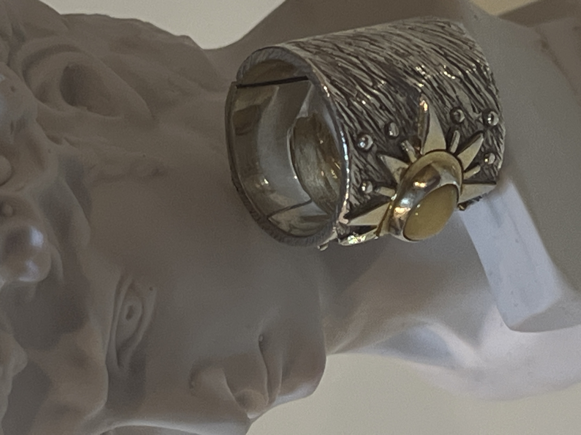 A close-up, eye-level, medium shot captures a sculpted bust, likely plaster, resembling a classical male figure, positioned at an indeterminate location in the city of Unknown, Unknown. The bust is light-colored, with defined facial features including an eye, nose, and mouth, though it appears incomplete, with the top of the head cut off by the frame. The focus is on the right side of the bust, where a wide, ornate ring is placed on the neck area of the sculpture. The ring is made of silver with a textured, striated band, and features a central, stylized gold sunburst design. Embedded within the sunburst is a single, oval, yellow gemstone. The lighting is soft and diffused, casting subtle shadows on the bust and highlighting the details of the ring.
