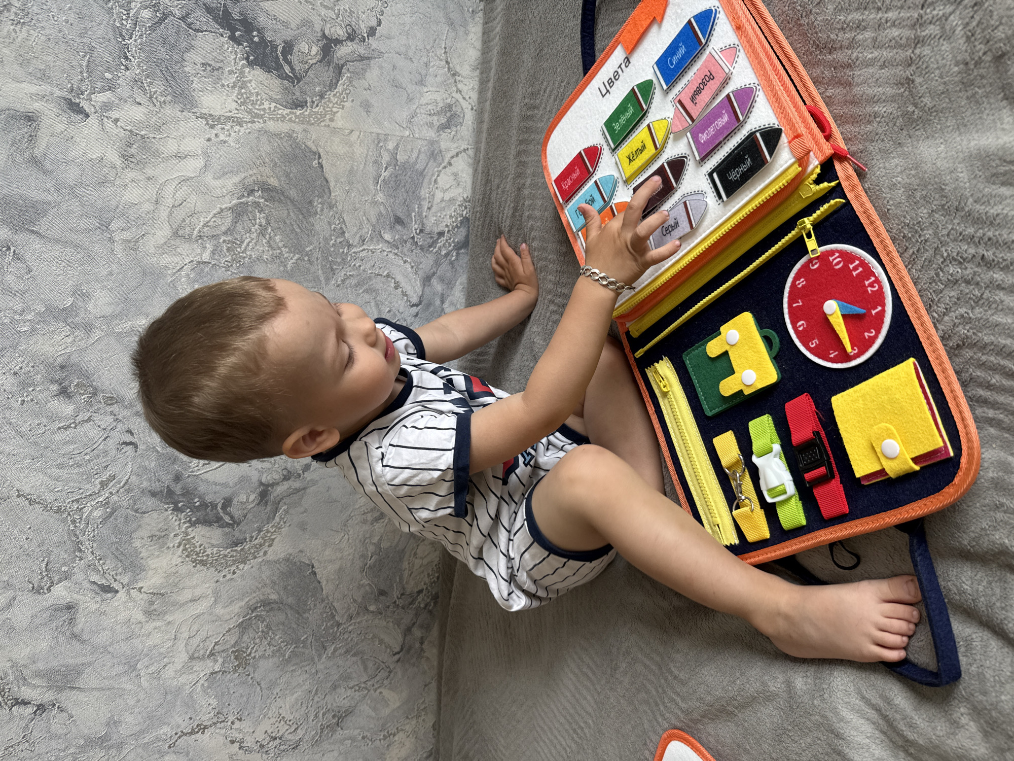A child is seated on a grey surface, possibly a bed or sofa, with their back against a textured, grey and white wall. They are wearing a striped, white and dark blue outfit and have a silver bracelet on their left wrist. The child is interacting with a colorful activity board with various elements such as color names written in Russian (Цвета), zippers, a clock face, and buckle straps. The focus of the image is on the child and their engagement with the activity board. The scene appears to be indoors. There is no indication that the image was taken in Rusanovo, Russia.