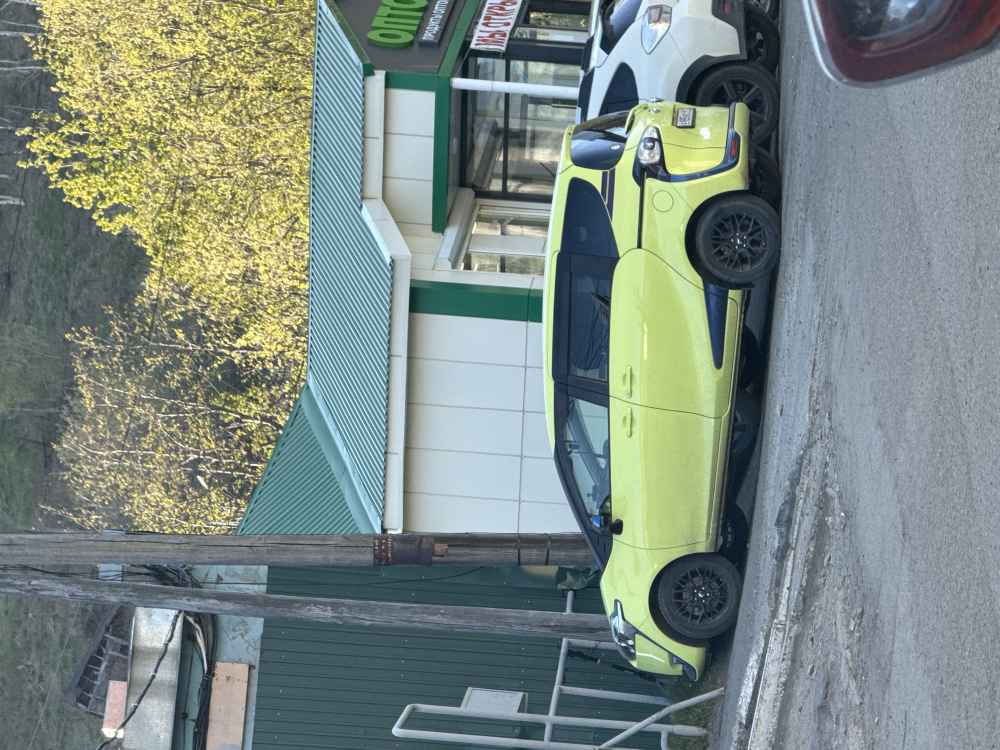 A lime green Toyota Sienta minivan, with navy blue trim, is parked along a curb next to a building with green and white trim. The building has a sign in Russian that says "OTTO" and "OTKPbl." A white car is parked behind the minivan. A utility pole with attached cables is in the foreground. Trees with yellow-green leaves are in the background. The street is paved. The scene appears to be in Petropavlovsk-Kamchatsky, Russia, based on the Russian signage.