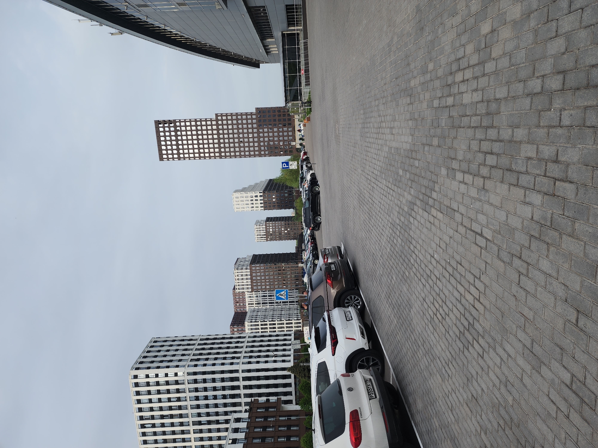 The image depicts a row of parked cars in Kazan, Russia, positioned along a paved area adjacent to a cluster of modern high-rise buildings. The buildings feature diverse architectural designs with repetitive window patterns. A parking sign and a pedestrian crossing sign are visible among the vehicles. The sky is overcast, suggesting a cloudy day. The scene appears urban and functional.
