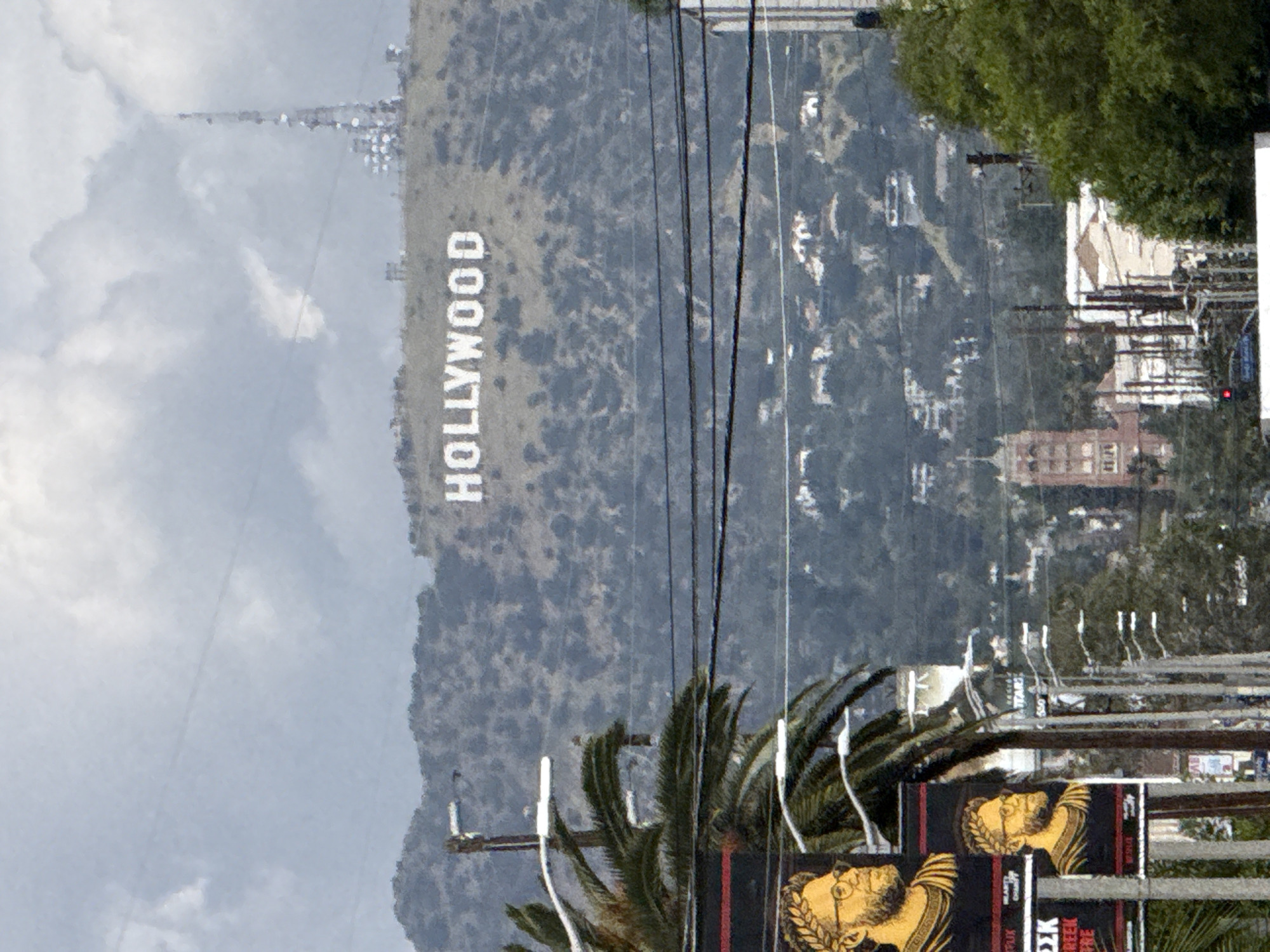 The image shows the Hollywood Sign, a landmark in the Hollywood Hills area of Los Angeles, California. The sign is positioned on the hillside against a backdrop of scattered trees and overcast sky. In the foreground, there is a billboard advertising "Greek Theatre" and some palm trees. Power lines are also visible cutting across the frame. A residential area with buildings is located in the middle ground.