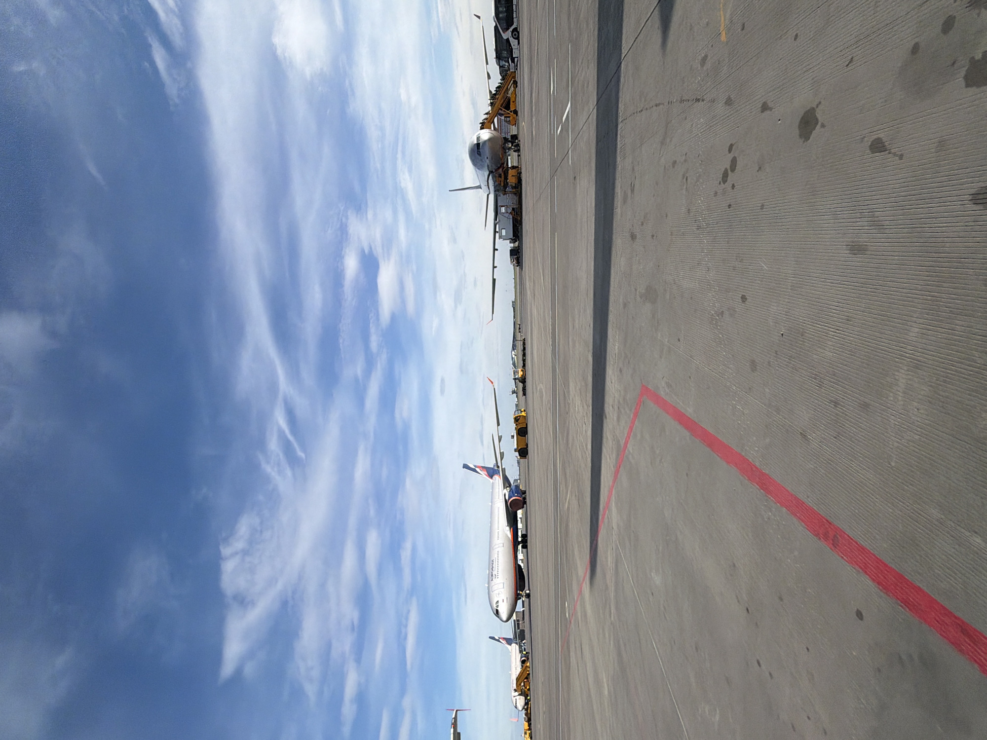 The image displays an active airport tarmac in Khimki, Russia, likely during the daytime under a bright, partly cloudy sky. The perspective is from the ground, looking across a wide expanse of concrete apron.
In the middle ground, a large passenger jet, identified as an "AEROFLOT" aircraft by its prominent text on the white fuselage and the distinct blue and red livery on its tail fin, is parked. This aircraft appears to be a wide-body, possibly an Airbus A330 or similar. Several pieces of yellow and white ground support equipment, such as passenger stairs and service vehicles, are visible around the aircraft, suggesting ongoing ground operations like loading, unloading, or maintenance preparation. The nose of another aircraft is partially visible further in the distance, hinting at a busy airport environment.
The concrete tarmac in the foreground and middle ground is textured with parallel grooves and features several dark spots, possibly oil or water stains. A prominent red painted line runs diagonally across the right side of the tarmac. The sky occupies the upper portion of the scene, showcasing varying shades of blue with scattered white clouds, ranging from wispy cirrus to more diffused formations. No people are clearly visible in the frame. The overall setting indicates a functional and operational airport environment.