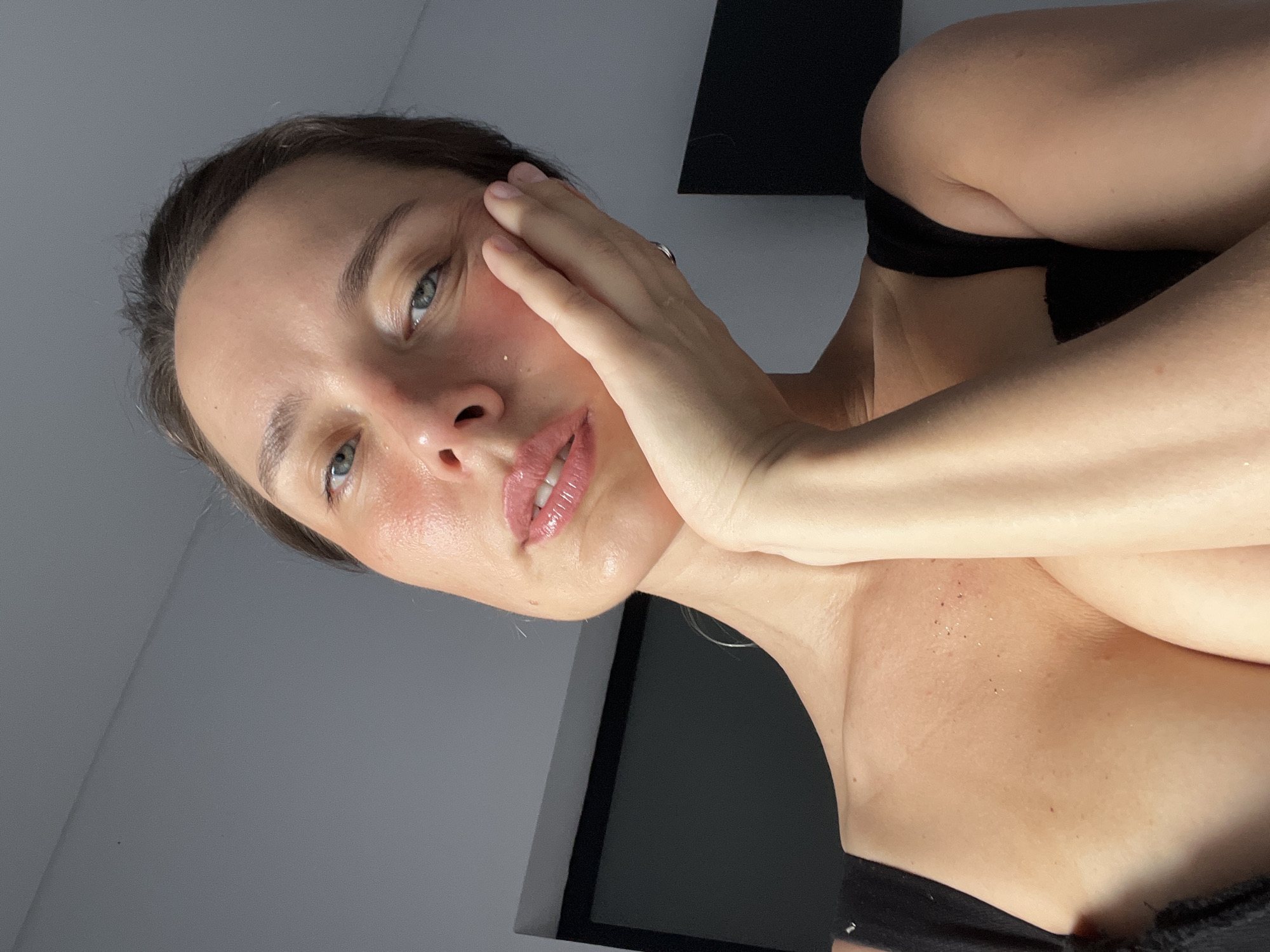 In North Kuta, Indonesia, a woman with light skin and light blue eyes, wearing a black top, poses for a selfie. She has her right hand resting on her cheek, with her fingers extended and her palm pressing against her face. Her lips are slightly parted, and she is looking towards the camera. The background is a plain gray wall.