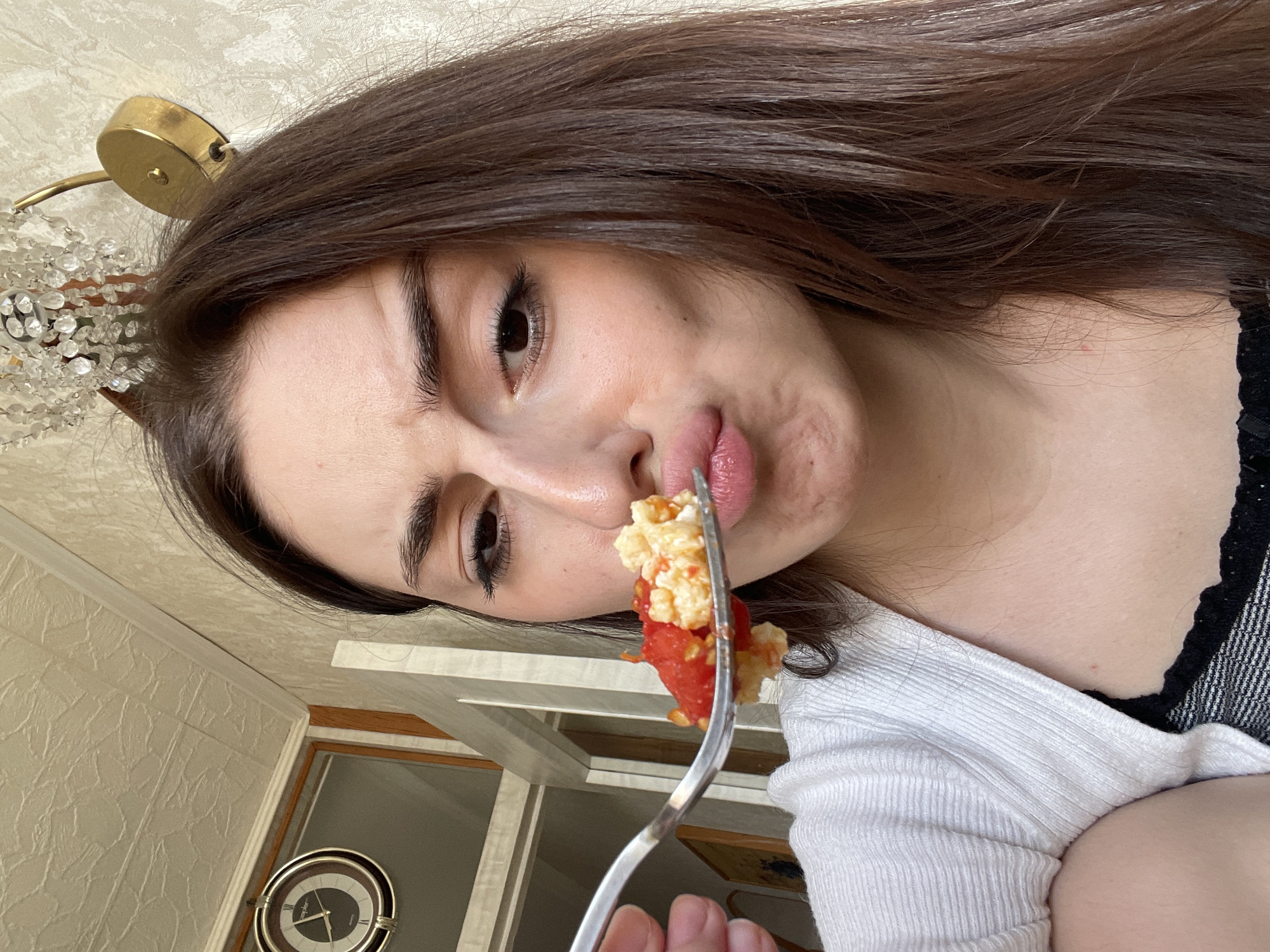 This image depicts a close-up, indoor scene of a young adult female about to consume food. The female, visible from the mid-torso up, has long, straight dark brown hair. She wears a white ribbed cardigan over a dark, possibly striped, garment with a lace trim. Her eyebrows are furrowed, and her lips are pursed as she looks at a silver fork held in her left hand, positioned near her mouth. The fork contains a portion of food consisting of a yellow/white granular substance mixed with red diced pieces, consistent with scrambled eggs and tomatoes.
The background is a residential interior in Moscow, Russia. To the upper left, a round analog wall clock with a dark face, gold-toned rim, and Roman numerals (X and II are partially visible) displays the time. Above the subject's head, a brass-colored wall-mounted light fixture or chandelier base supports multiple clear crystal pendants. The walls are covered in a light-colored, textured wallpaper. The ceiling also features a textured pattern. A window or doorway with light wood trim is partially visible in the left background, allowing for natural light into the space. The specific address or neighborhood within Moscow cannot be determined from the visual information provided.