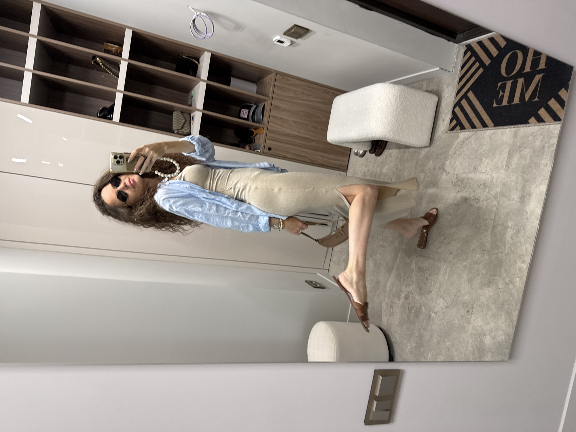 A woman stands in front of a full-length mirror in what appears to be a closet or dressing room, taking a selfie with her smartphone. She has long, dark, curly hair and is wearing sunglasses, a light blue unbuttoned shirt over a beige fitted dress with a thigh-high slit, a pearl necklace, and brown flat sandals. She is holding a small, tan shoulder bag in her left hand and has her right leg kicked out behind her, with her right foot pointed. In the background, to her right, is a tall wooden shelving unit filled with handbags, hats, and other accessories. To her left and behind her is a round, off-white upholstered ottoman. The floor is tiled with a marble-like pattern, and a black doormat with tan geometric patterns and the word "HOME" is visible in the lower right corner. The walls are a neutral light gray.
