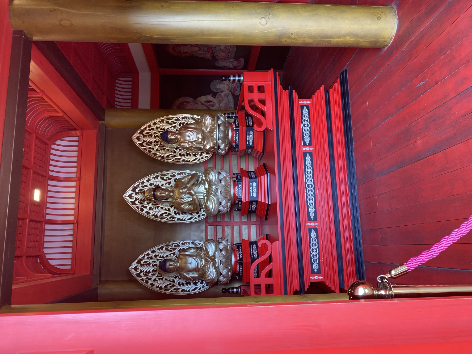 This image captures an interior scene, likely a shrine or exhibit, characterized by a dominant red and gold color palette. Three golden statues, resembling seated Buddhist figures, are centrally positioned on a multi-tiered red and black altar-like structure. Each statue features a blue topknot/hair, golden skin, and distinct hand gestures (mudras), with a golden, ornate, flame-shaped mandorla serving as a backdrop. The statues are set upon individual red and black pedestals adorned with silver and white decorative patterns, which rest on the larger red base.
The flooring consists of red-stained wooden planks. To the right, a large, cylindrical, golden-colored pillar extends from floor to ceiling. The wall behind the statues is a plain dark brown. Further to the right, beyond the golden pillar, dark walls display murals or tapestries with lighter-toned depictions of seated figures, possibly deities. The left wall and the visible doorway frame are a vibrant red. The ceiling is intricately designed with red wooden latticework, arched sections, and white recessed panels, with a single light fixture visible. In the lower left foreground, a polished brass stanchion with a spherical top and a bright pink braided rope acts as a barrier. This interior scene is located in Krasnodar, Russia.