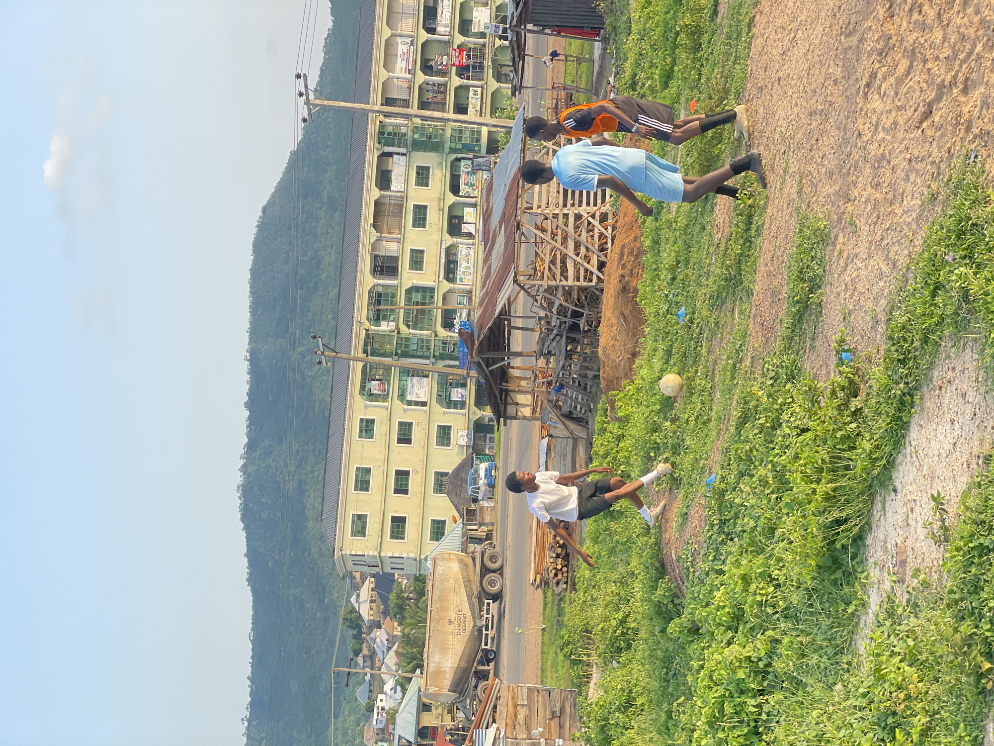 In Lokoja, Nigeria, three young males are engaged in a game of soccer on a dirt field surrounded by lush green vegetation. A large, light-colored cement truck is parked on the adjacent road. Behind the players, a multi-story building with a light green facade stands prominently, featuring numerous windows and balconies. To the right of the building, a smaller, simpler structure is visible. The background is dominated by a gently sloping, tree-covered hill under a clear, pale blue sky with a few wispy clouds. The overall scene depicts a casual, outdoor recreational activity within a semi-urban environment.