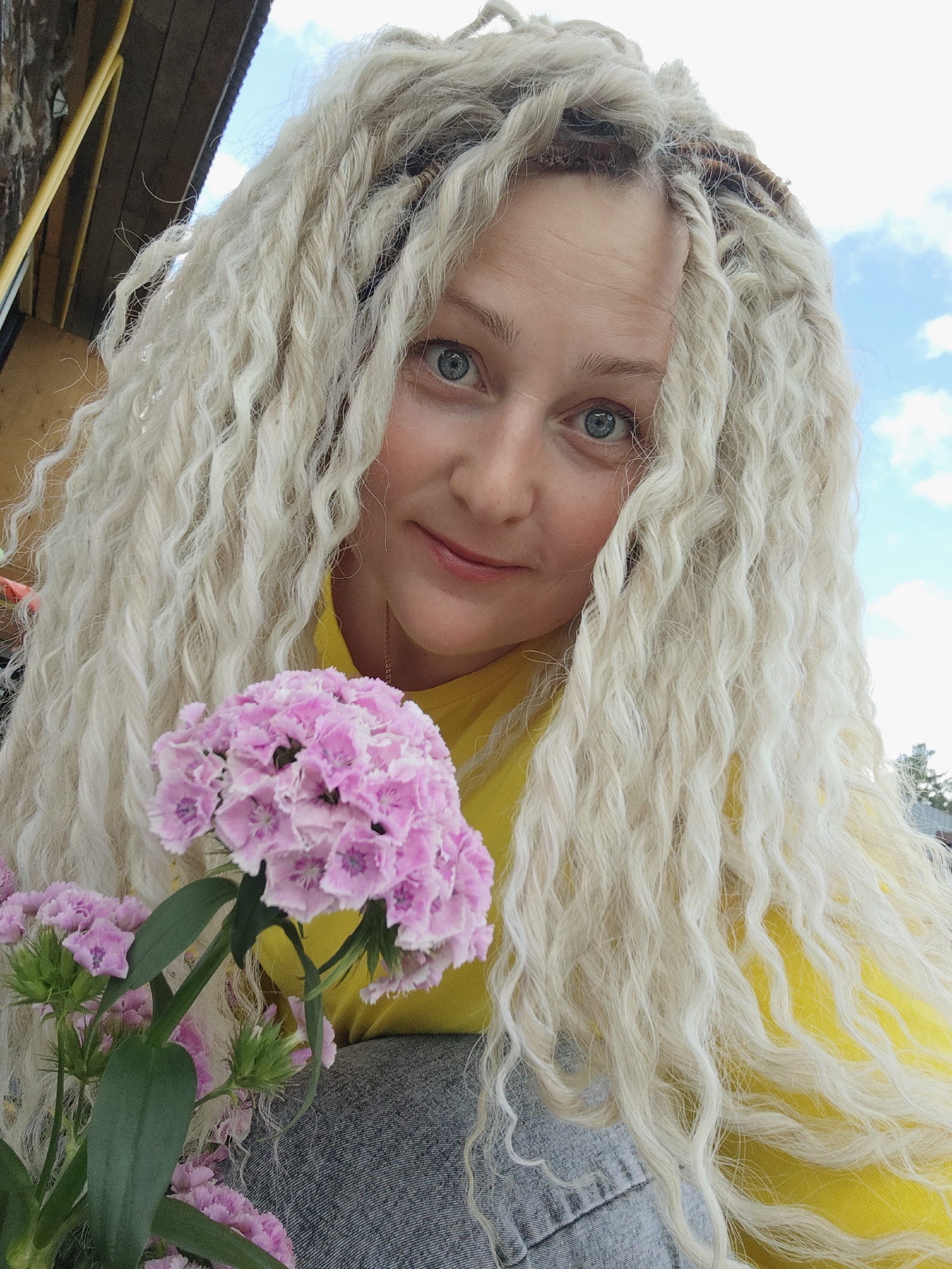 A young woman with long, blonde dreadlocks and blue eyes is holding a small bouquet of pink flowers. She is wearing a yellow shirt and a grey denim jacket. The woman is smiling and looking at the camera, with the sky and some wooden structures in the background. The image appears to have been taken outdoors on a sunny day. There is no visible text in the image. The woman's expression is happy and serene.