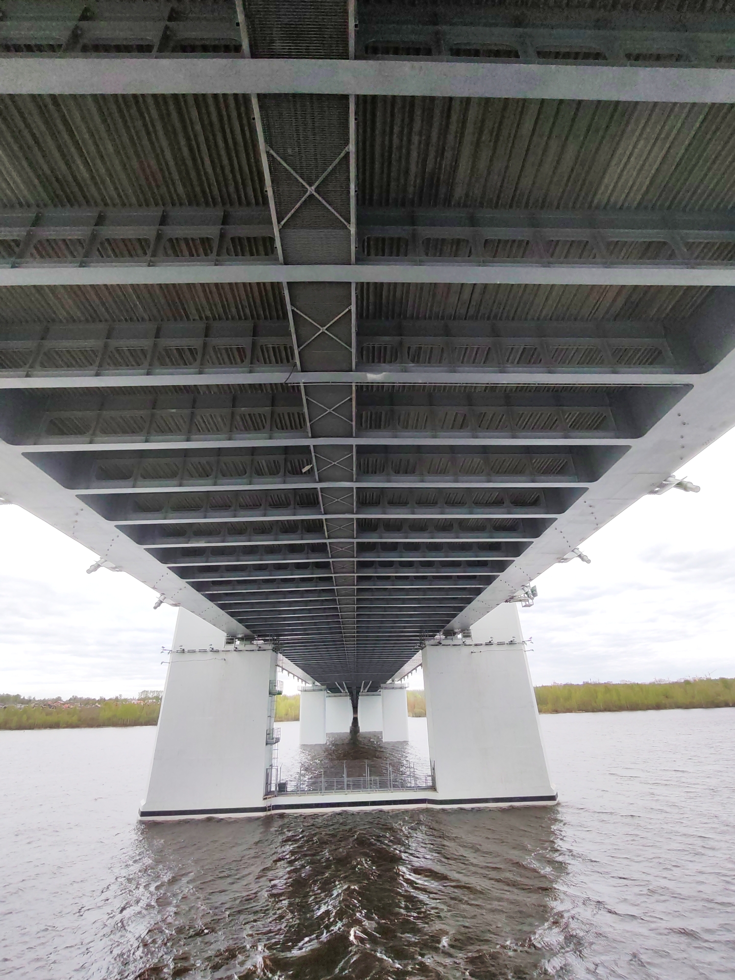 The image shows the underside of the Oktyabrsky Bridge in Cherepovets, Russia. The bridge is constructed of gray metal beams and supports resting on white concrete pillars that descend into the water. The water surface is rippled and reflects the gray sky. In the background, there is a strip of green vegetation on the shoreline.