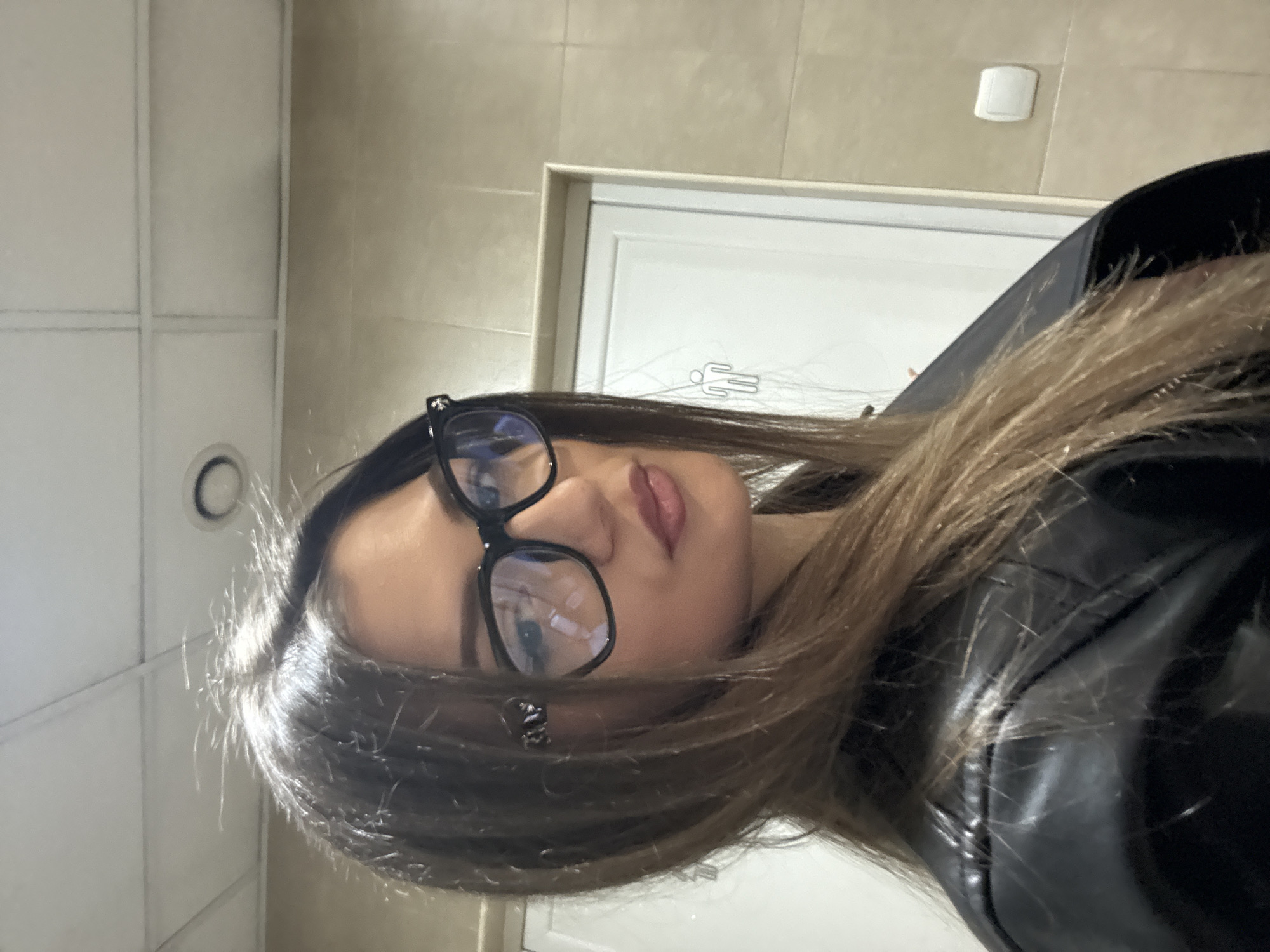The image is a selfie of a woman wearing glasses and a black leather jacket, taken in what appears to be a public restroom. The background features tiled walls, a white door with a stick-figure sign typically indicating restroom facilities, and a small rectangular sensor, likely for an automatic light or soap dispenser. There is no specific information to suggest the location is in Sofia, Bulgaria.
