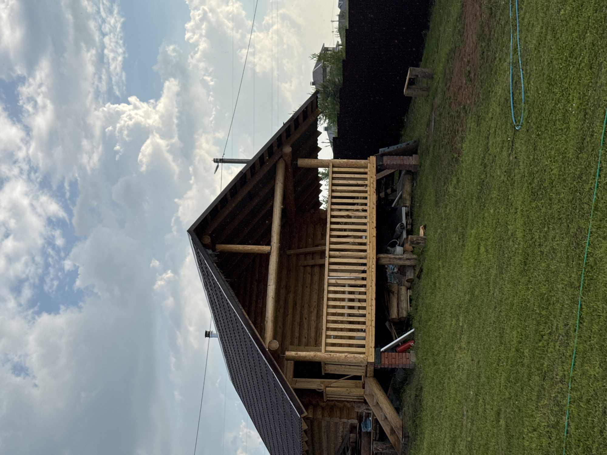 A log cabin with a dark metal roof is situated on a grassy incline under a cloudy sky. The cabin features a wooden porch with a railing and stairs leading up to it. The porch is supported by brick pillars and wooden posts. To the left of the cabin, there are stacks of lumber and other building materials. A dark fence runs along the right side of the property, with some trees and houses visible in the distance. The foreground is a well-maintained lawn with a blue hose laid out on the grass. The image appears to be taken in a rural or suburban setting, potentially a dacha or a private residence. The specific location in Izhevsk, Russia, is not identifiable from the visual information provided.