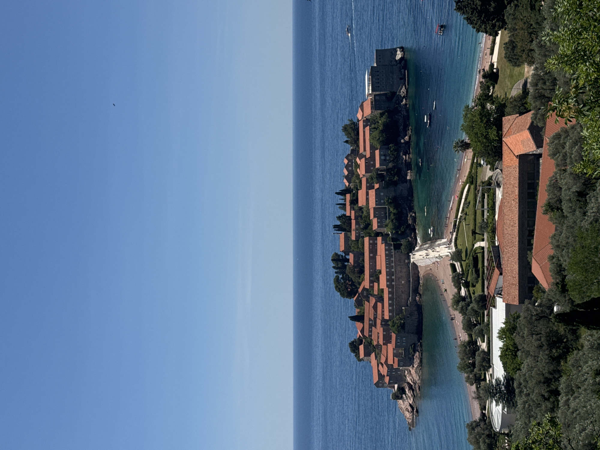 The image shows Sveti Stefan, Montenegro, an island resort connected to the mainland by a narrow isthmus. The island is densely packed with stone buildings, mostly with terracotta-colored roofs. The surrounding sea is a deep blue, with boats visible in the distance. A sandy beach stretches along the coastline next to the island. Adjacent to the beach is a hotel complex with terracotta-roofed buildings and manicured gardens. The scene is viewed from an elevated perspective, with trees and foliage framing the right side of the image. The sky is a clear blue.