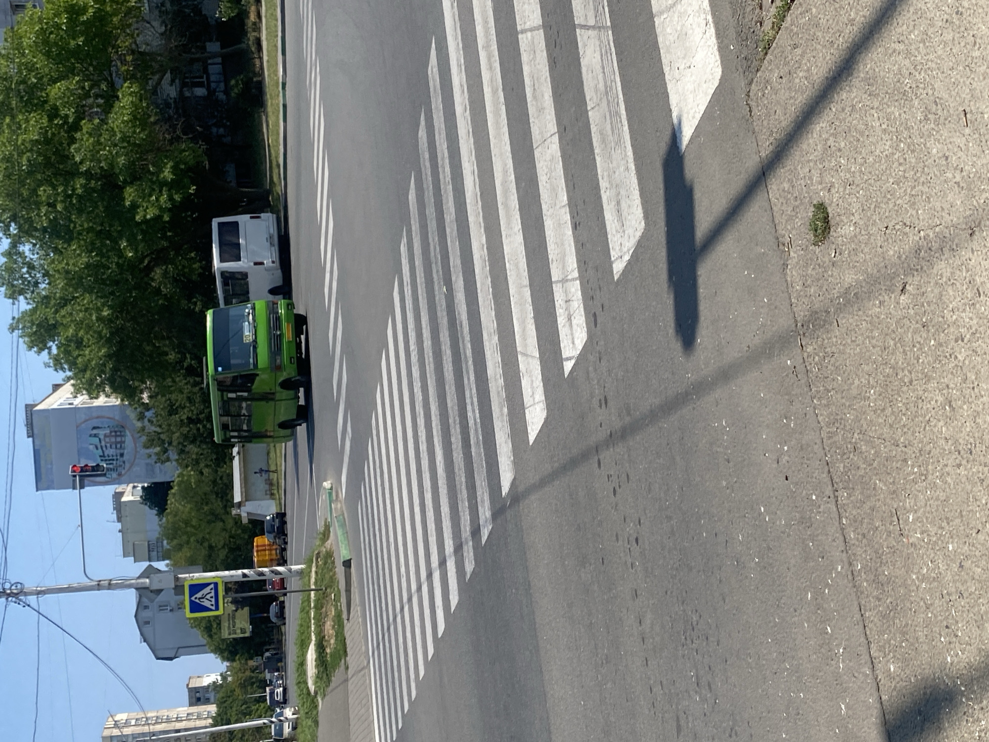 The image depicts a street scene in Chornomorsk, Ukraine, on a sunny day. A prominent pedestrian crossing with white stripes is visible in the foreground. To the left, a traffic light is red. Adjacent to the traffic light is a blue and yellow pedestrian crossing sign. A bright green minibus is positioned at the crossing, and a white bus is behind it. In the background, buildings and lush green trees line the street. The overall impression is a typical urban intersection with ongoing traffic.