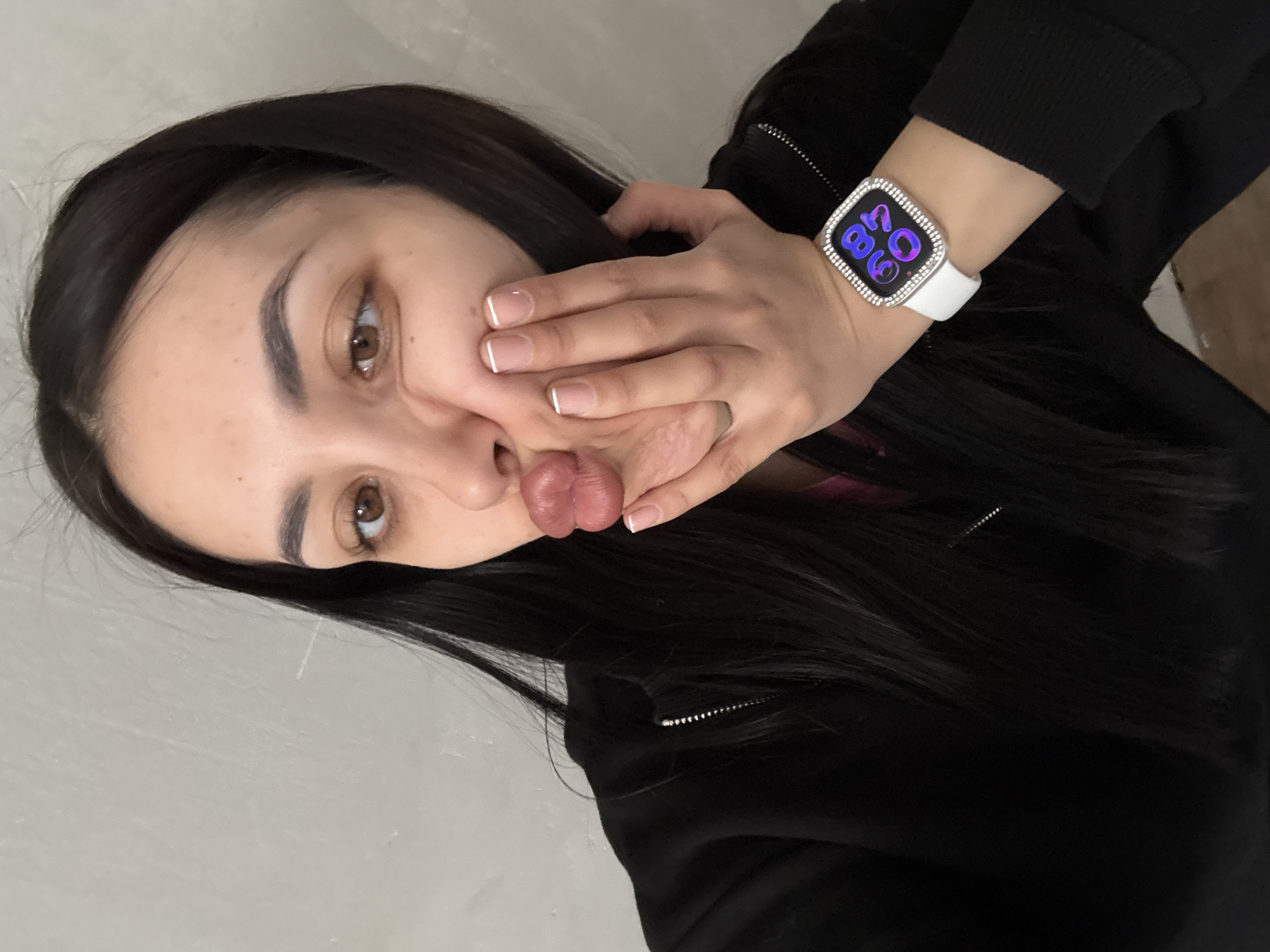 A woman with long dark hair, brown eyes, and manicured nails is posing for a selfie. She is puckering her lips. Her right hand is touching her face, and her left wrist is visible, displaying a white smartwatch. She is wearing a black zip-up jacket. The background appears to be a light gray wall. There is no indication of Votkinsk, Russia in the image.