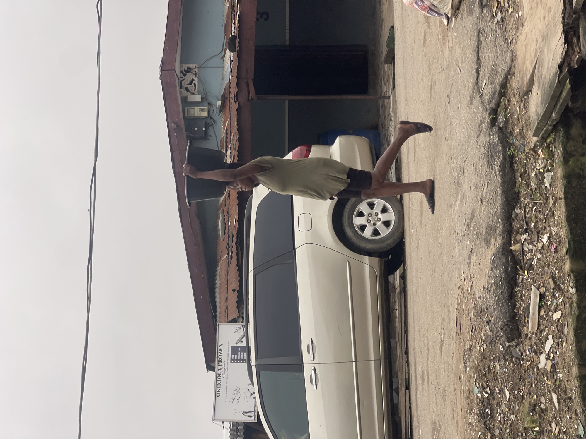 A woman is walking from left to right across a paved street in Akowonjo, Nigeria, carrying a large black tub on her head. She is wearing a loose-fitting olive green t-shirt and black shorts. A beige minivan is parked on the left side of the street, partially obscuring the woman. In the background, a light blue building with a corrugated metal roof is visible, with a sign above it that reads "OKIKiola FROZEN Sea Foods." The street is unkempt, with debris scattered on the ground, and the pavement is cracked and uneven. The sky is overcast.