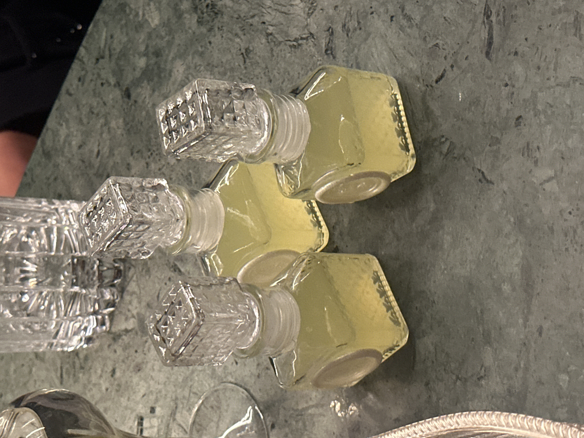 The scene depicts a still life arrangement on a dark marble or granite surface. Three small, square decanters filled with a yellow liquid are positioned in a staggered line. Each decanter has a clear, ornate, cubic stopper. Beside the decanters is a larger, similarly ornate decanter, but with more intricate cut glass designs. The context suggests an interior setting, possibly a dining room or bar. There are no indications that the location is specifically in the city of London, United Kingdom.