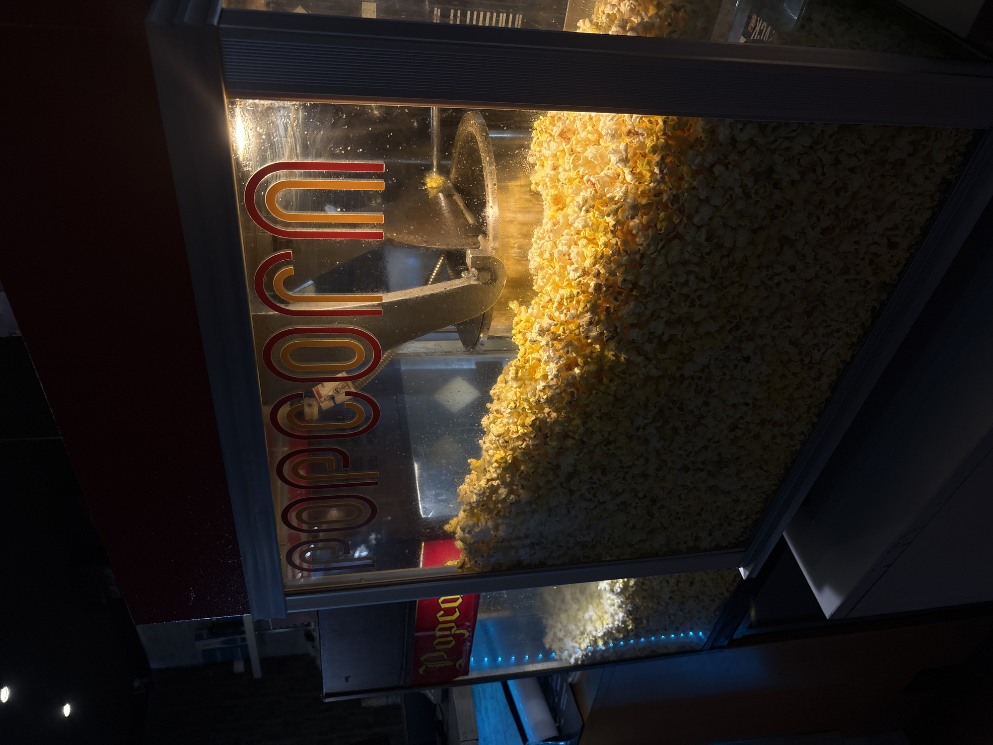 An eye-level shot depicts a popcorn machine filled with popcorn. A rotating metal component inside the machine is visible, partially obscured by the popcorn. The word "Popcorn" is emblazoned on the front of the machine. The environment is dimly lit. Location and information about Gotse Delchev, Bulgaria are not discernible.