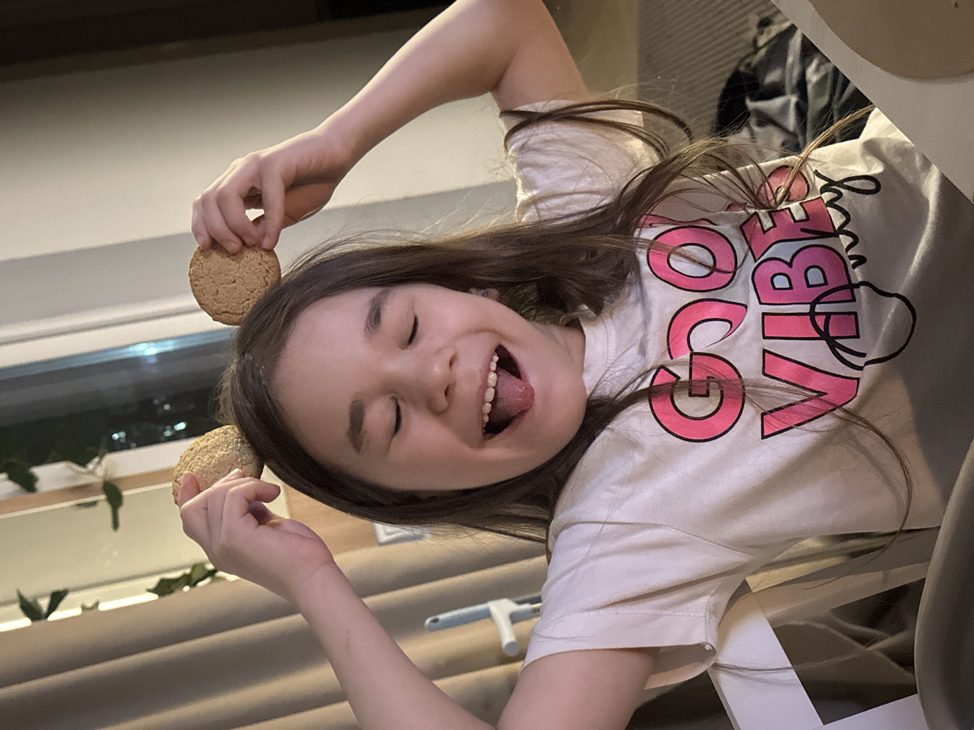 A young girl with brown hair and a white t-shirt with the words "GOOD VIBES" in pink lettering, is holding two oatmeal cookies up to her head while sticking her tongue out, smiling, and looking off to the side. She is in an indoor environment with blinds and a window in the background. The exact location in Kharnavy, Russia, is not identifiable.