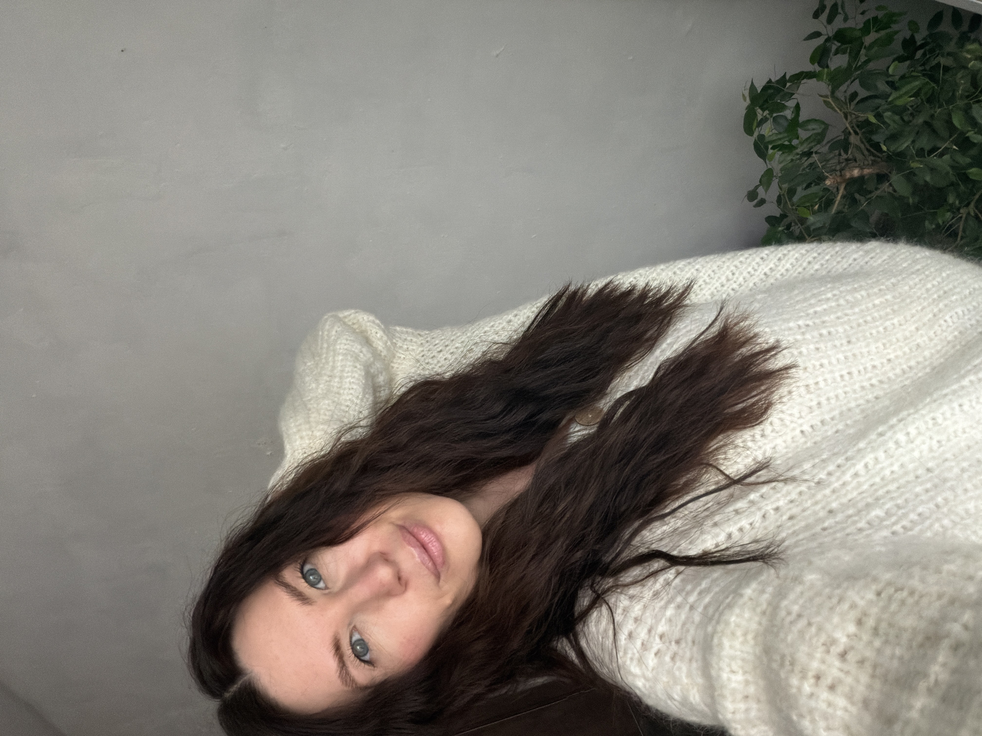 A female subject, appearing to be in her late 20s to early 40s, is depicted in a medium-close portrait shot. She has fair skin, blue eyes, and long, wavy dark brown hair parted centrally. Her lips are light pink, and she exhibits a slight smile while looking directly at the viewer. She is wearing a cream-colored, chunky knit sweater with a visible light brown button near the collarbone. Her left arm is raised, with the hand positioned behind her head or upper neck. The background consists of a plain, light grey, possibly textured wall. In the lower right corner, a portion of a houseplant with dark green leaves and visible branches is present, partially obscured by the subject's sweater. The lighting is soft and even, consistent with an indoor environment. The specific location within Perm, Russia, is not discernible from the provided image.
