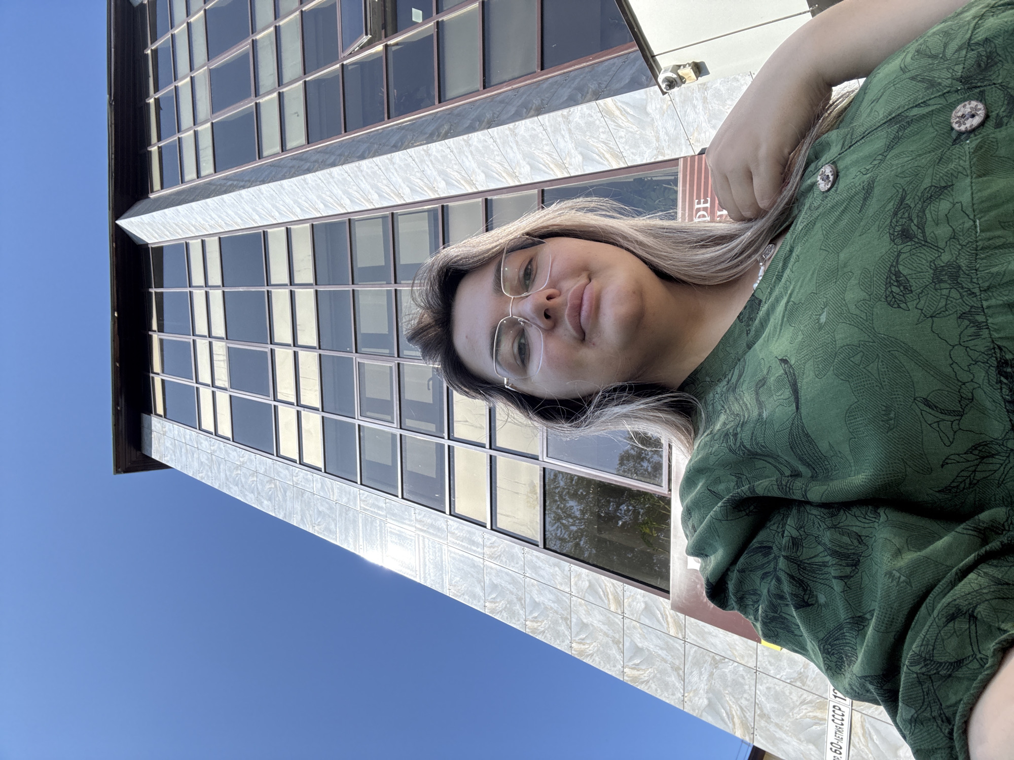 A young woman with blonde hair and glasses takes a selfie in front of a tall, modern building. The building has a facade of marble and glass windows, and it is set against a clear blue sky. The woman is wearing a green, patterned dress and a necklace. A portion of a sign with Cyrillic text, "пр. 60-ЛЕТИЯ СССР, 17" (which translates to "60th Anniversary of the USSR Avenue, 17"), is visible on the building, indicating the location is Birobidzhan, Russia. The image is taken from a low angle, looking up at the building and the woman.