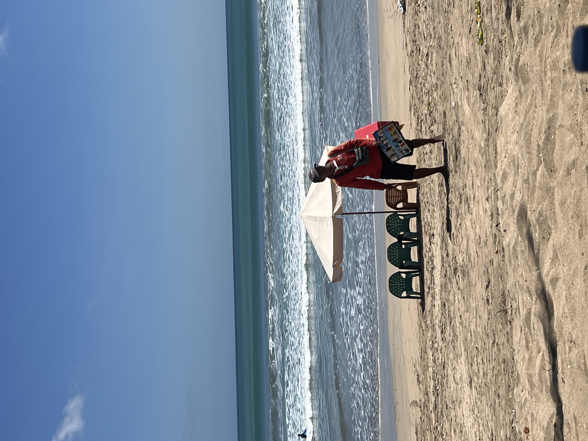 On a sunny day at Kuta Beach, Indonesia, a vendor wearing a red long-sleeved shirt, black shorts, a baseball cap, and a crossbody bag walks along the sandy shore. He carries a red cooler bag in his right arm and a flat, rectangular object with colorful pictures in his left. In the background, the turquoise ocean meets the horizon under a clear blue sky with a few wispy clouds. Gentle waves break on the shore, creating a white foamy edge. Further down the beach, a beige umbrella shades four green plastic chairs and one brown plastic chair. A lone surfer is visible in the distance, riding a wave. The sand is marked with footprints and scattered debris.
