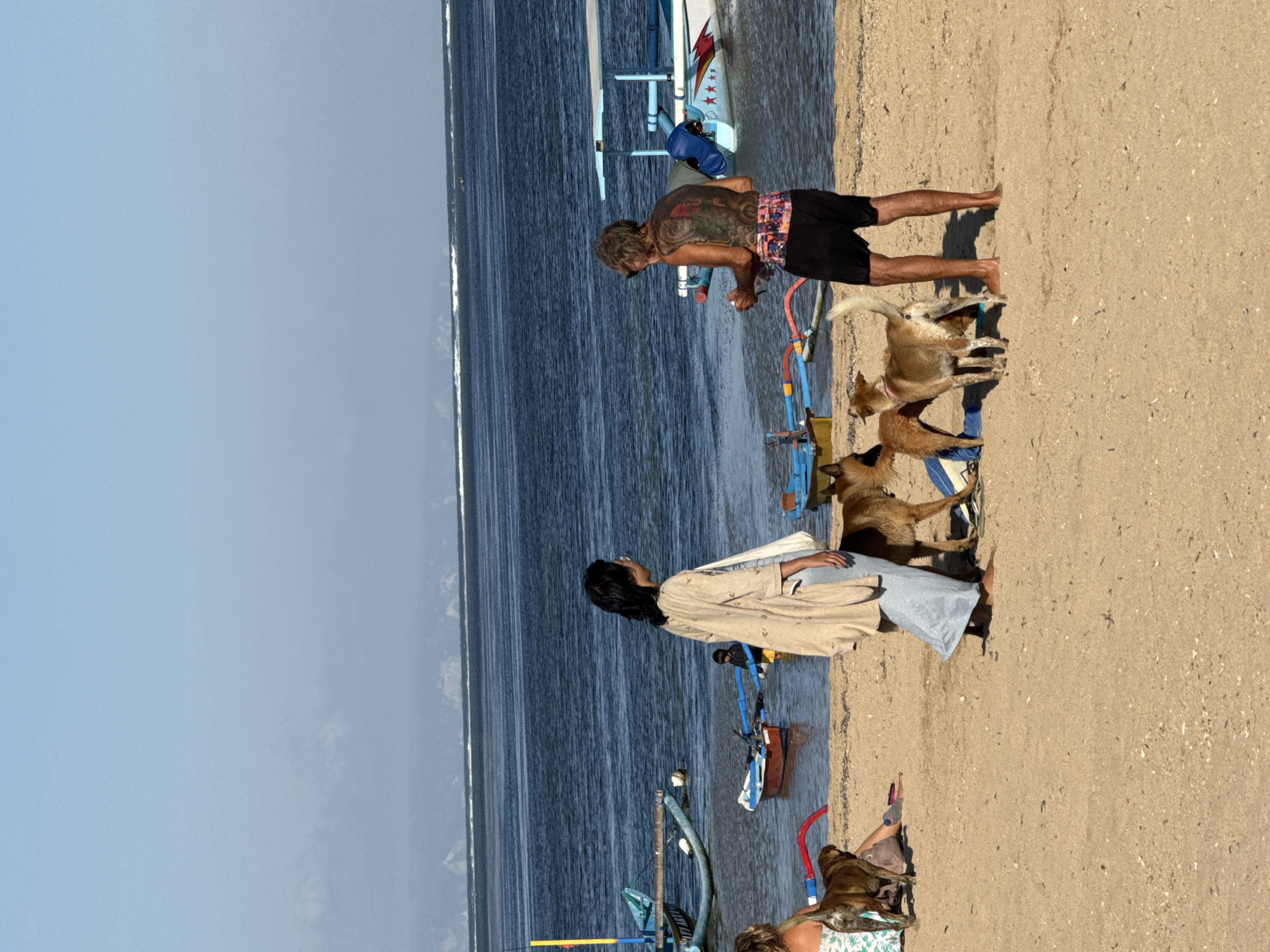 The image depicts a sunny day at a beach in Denpasar, Indonesia. The foreground shows a sandy beach with several people and dogs. A woman in a light-colored robe and grey skirt is standing near the water's edge, facing away from the camera. A man with extensive tattoos on his back, wearing black shorts, is standing a short distance away, also facing away. Two dogs are interacting with the man, and another dog is visible near a seated person on the left side of the frame. In the background, the ocean is calm, with a few traditional Balinese fishing boats (jukung) anchored near the shore. The horizon shows a hazy outline of land. The overall scene conveys a relaxed atmosphere by the sea.