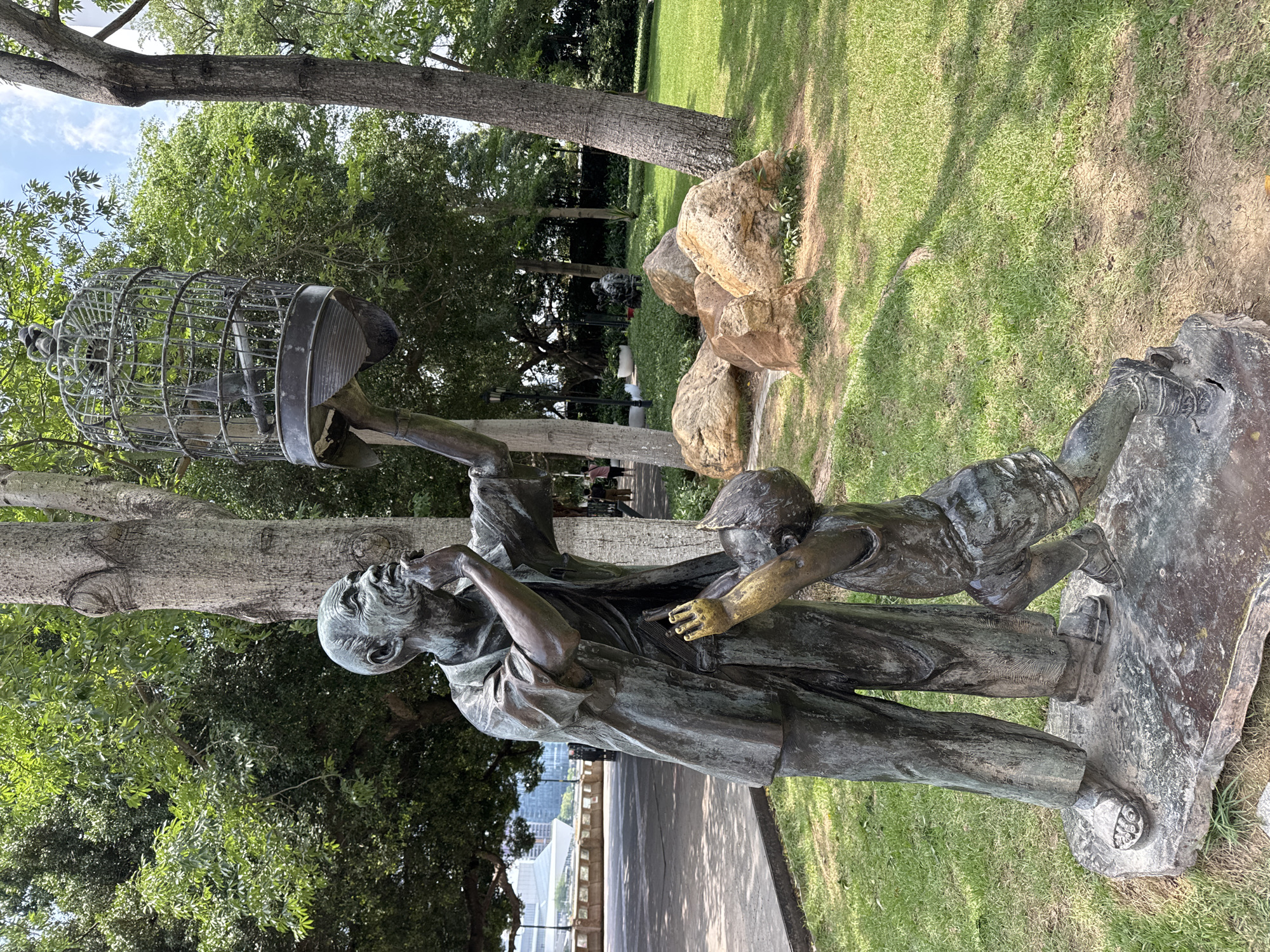 A bronze sculpture in a park setting depicts an elderly man holding a birdcage aloft while a young boy reaches into it. The sculpture is positioned near a walkway and overlooks a cityscape. The scene includes trees, grass, and large rocks. The sculpture is located in the city of Liwan District, China.
