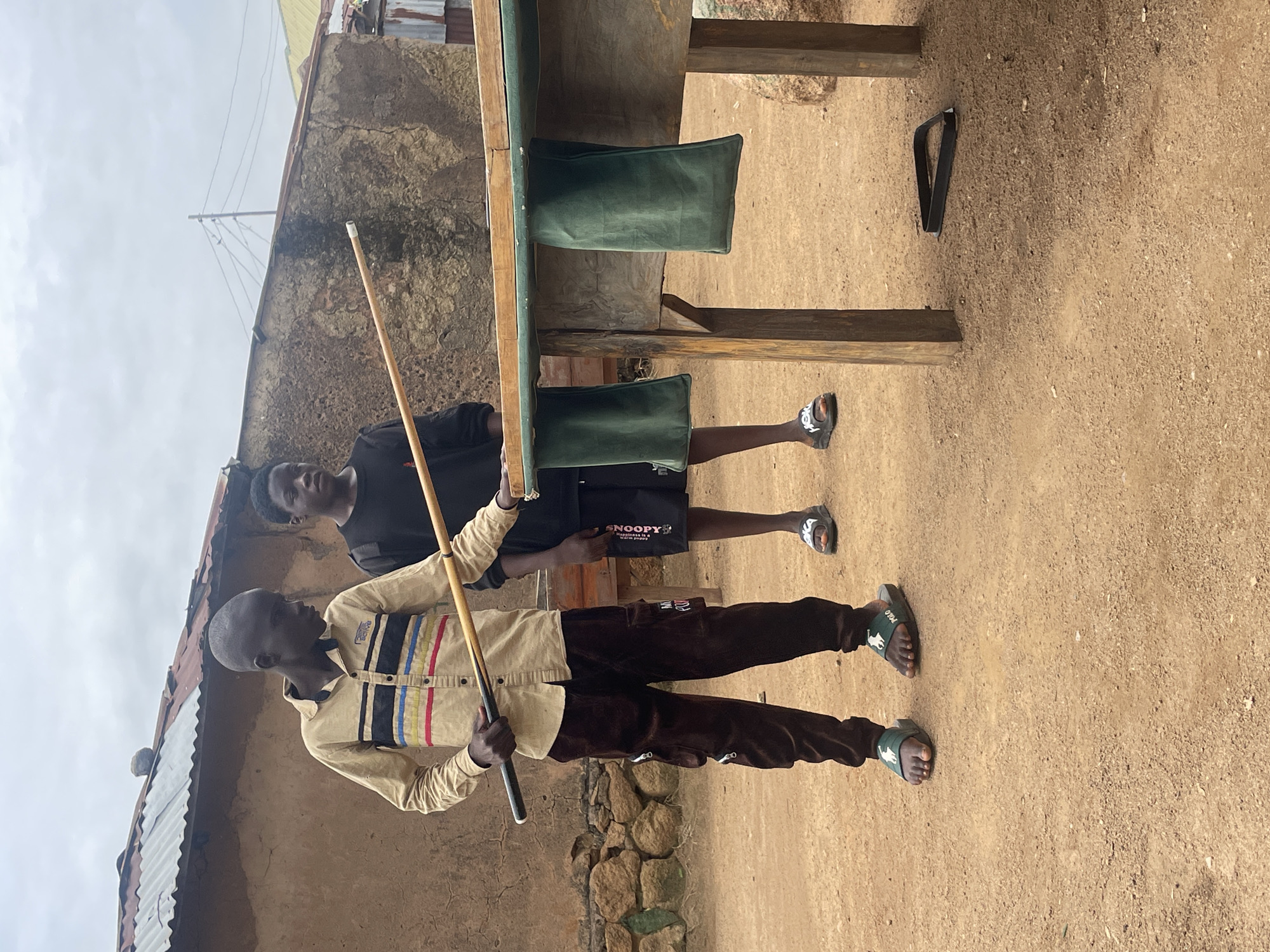 Two young males, appearing to be teenagers, are standing near a pool table outdoors, likely in a residential area of Jos, Nigeria. The male on the left, wearing a tan and striped long-sleeved shirt and brown cargo pants, holds a pool cue as if preparing to take a shot. The male on the right, dressed in a black t-shirt and black shorts with white lettering, stands beside him, observing. The pool table, with green felt pockets, is positioned on wooden legs. The ground is a reddish-brown dirt surface, typical of the area. In the background, a simple building constructed from mud bricks with a corrugated metal roof is visible, along with some scattered rocks and vegetation. The sky is overcast. The overall scene suggests a casual outdoor game being played in a community setting.
