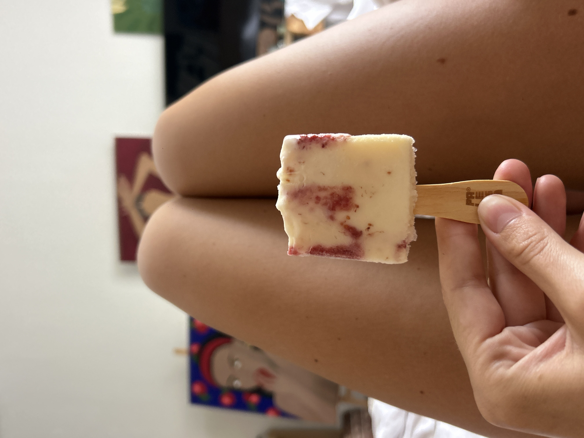 A person holds a white popsicle with red swirls on a wooden stick in their right hand. The background is blurred, but shows fair skin, two paintings, and a black object. It cannot be determined if the image was taken in North Kuta, Indonesia.