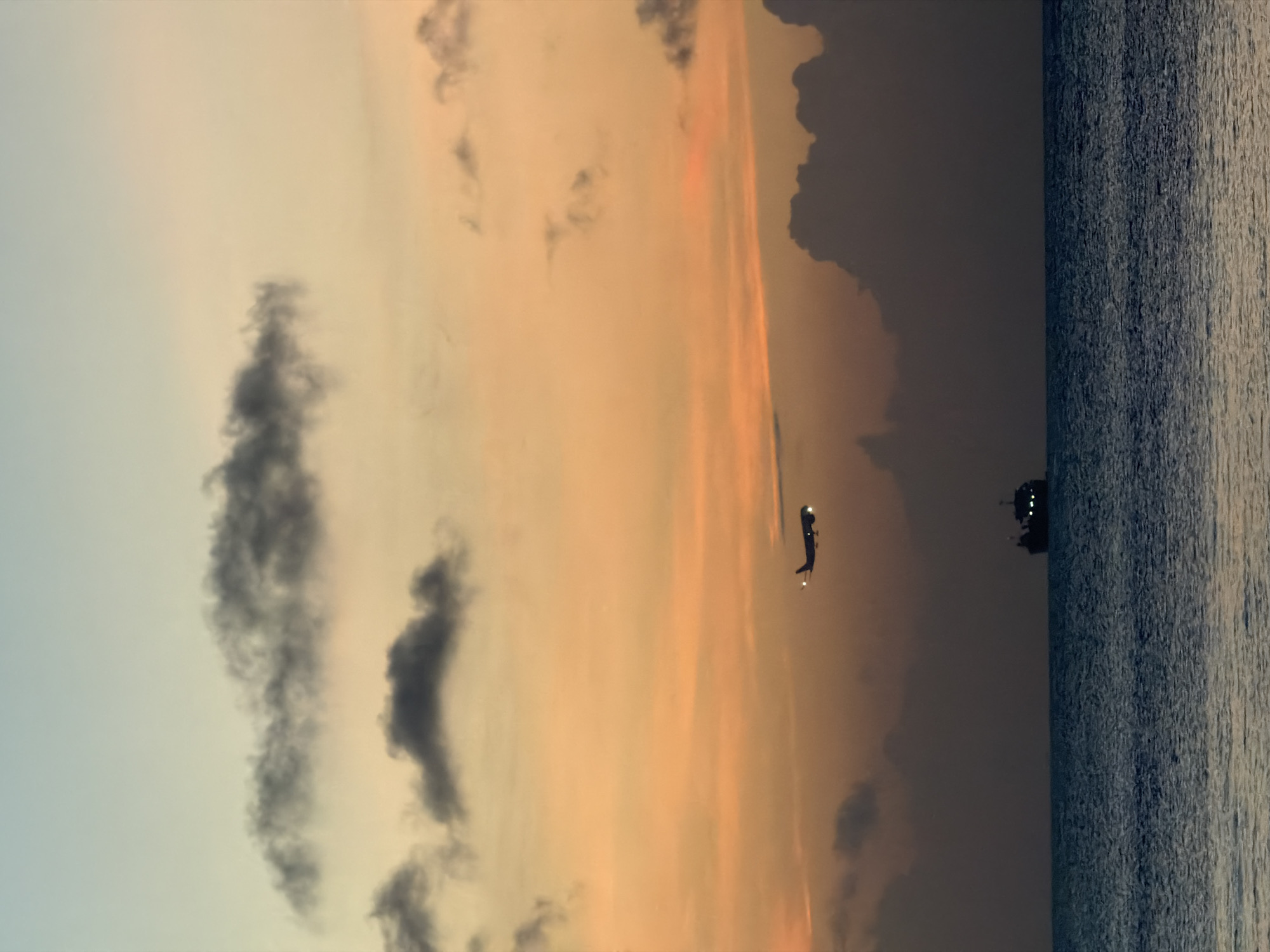 A horizontally-oriented image captures a sunset scene. The right side of the image shows the ocean with water lines, and the left side shows the sky filled with clouds. The sky transitions in color from orange and red hues near the center to a lighter blue color on the far left. The clouds vary in size and color from dark gray to white. A plane is visible in the center of the image. According to geolocation information, the location is Kuta Selatan, Indonesia.