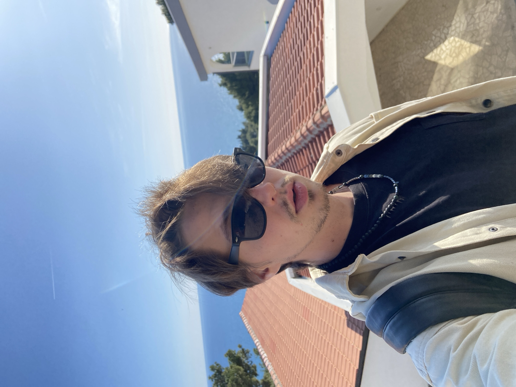 A person with light brown hair and sunglasses is facing the camera. They are wearing a black t-shirt and a light-colored jacket. A beaded necklace is visible around their neck. The background includes the sky, the sea, trees, and buildings with red tiled roofs, likely indicating a location in Šušanj, Montenegro. The angle of the photograph suggests it was taken from an elevated position.