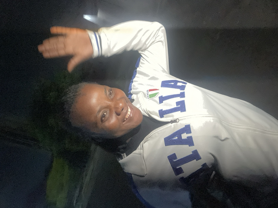 A woman is smiling and waving while wearing a white jacket with "ITALIA" written in blue letters. The Italian flag is visible above the lettering. The background is dark and out of focus, suggesting a nighttime setting. The lighting is uneven, with strong highlights on the jacket and the woman's face. There appears to be greenery on the left side of the image.