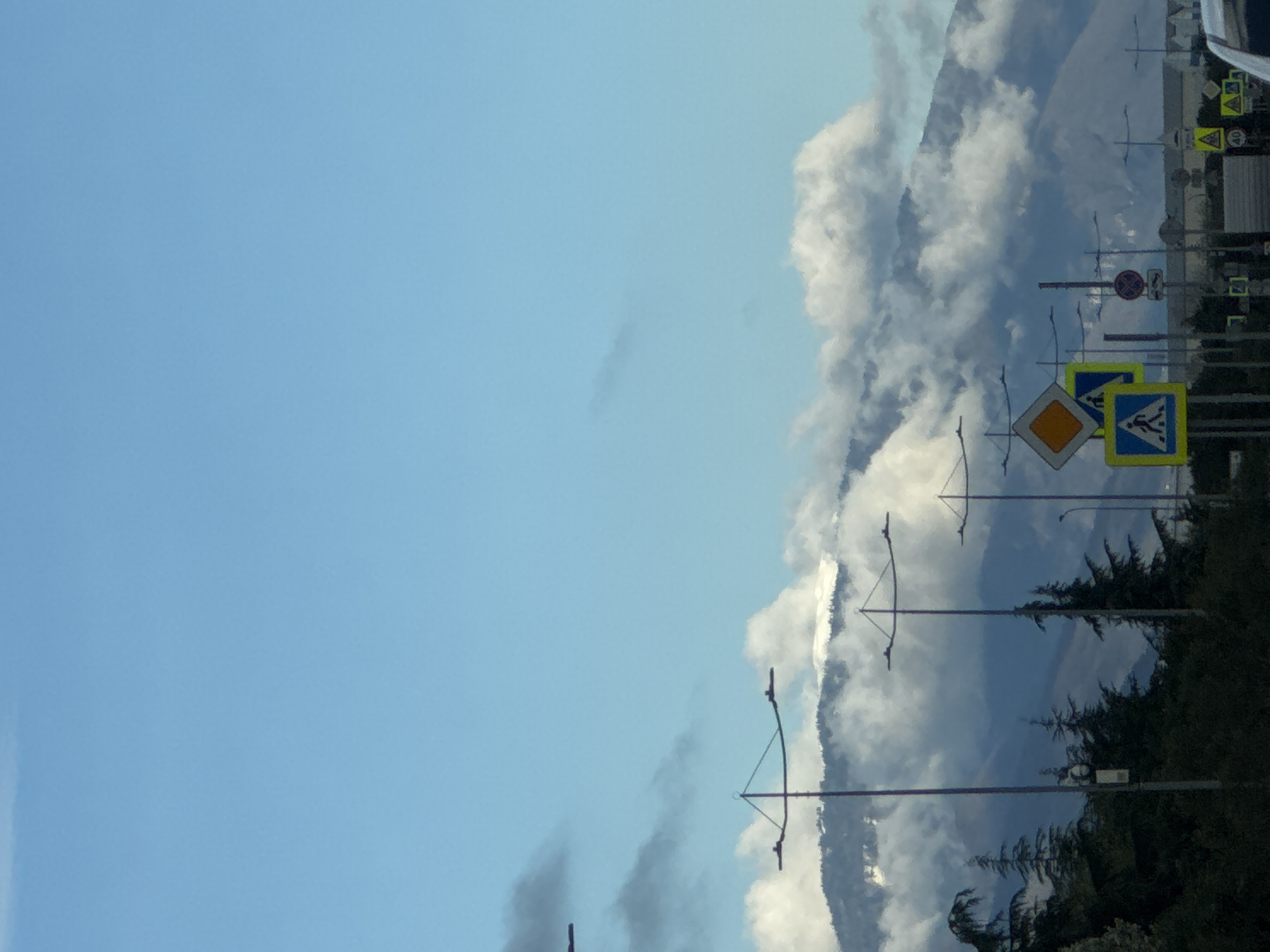 The image presents a scene from Sochi, Russia. The sky is a bright blue, with white clouds scattered towards the right of the frame. The right side features roadside infrastructure, including traffic signs, lampposts, and trees. The signs indicate pedestrian crossings and speed limits. The background suggests a mountainous landscape obscured by clouds.