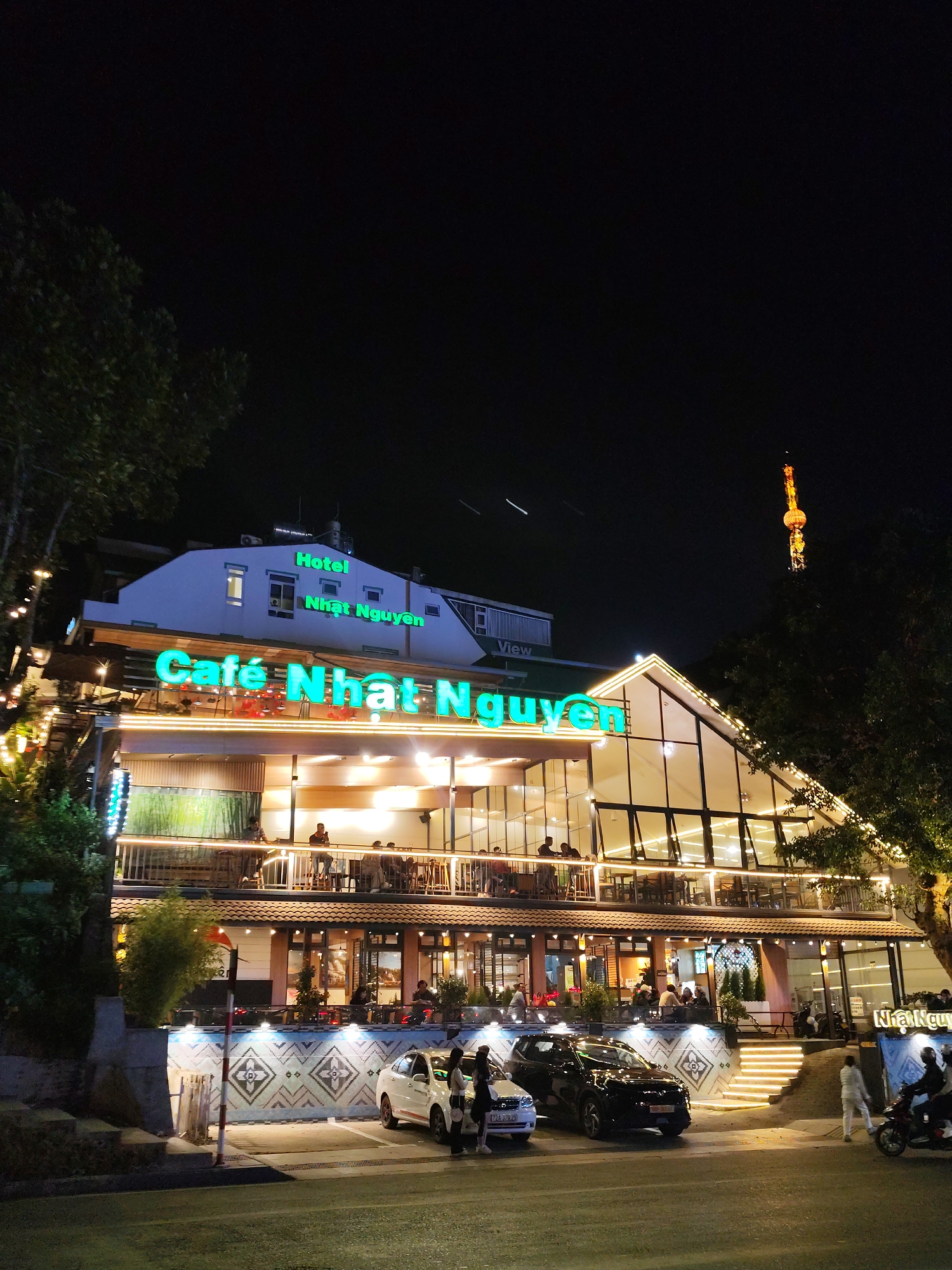 The image shows a multi-story building at night identified as "Cafe Nhat Nguyen" and "Hotel Nhat Nguyen" in Da Lat, Vietnam. The buildings are illuminated with bright lights, with several people visible on the balconies and inside the establishment. Two cars are parked in front of the building, and a motorcycle is visible on the right side of the frame. A tower-like structure with decorative lighting is seen in the background. The overall atmosphere is lively and urban, with a focus on the architecture and the activity surrounding the cafe and hotel.