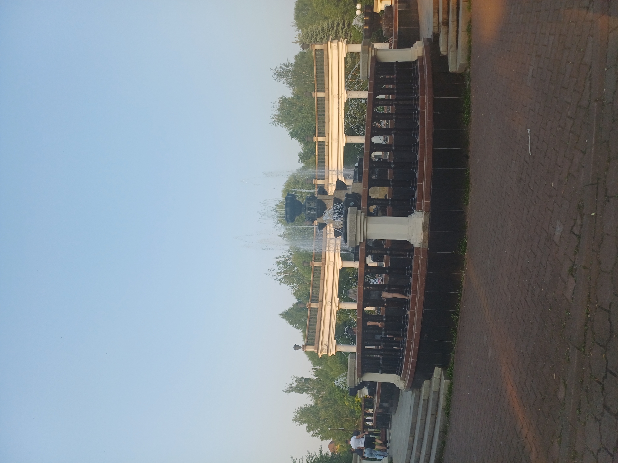 The image shows an outdoor scene with a multi-tiered fountain in the foreground, set against a backdrop of trees and a clear sky. The fountain is ornate, featuring dark urns and water jets that create a spray. It's situated along a curved walkway made of brick pavers.
A light-colored railing with dark vertical supports lines the fountain's edge. Behind the railing, there appears to be a walkway or structure with columns. People are visible in the scene, some walking along the paved area, others standing near the fountain. One group of people includes at least two individuals wearing white shirts and blue jeans.
The time of day appears to be late afternoon or early evening, as indicated by the warm, golden light illuminating parts of the scene, particularly the brick walkway and the upper structures. The weather seems pleasant, with a clear, light blue sky. The overall setting suggests a public park or promenade. There is no visible text in the image. The composition and the presence of people suggest a leisurely atmosphere.