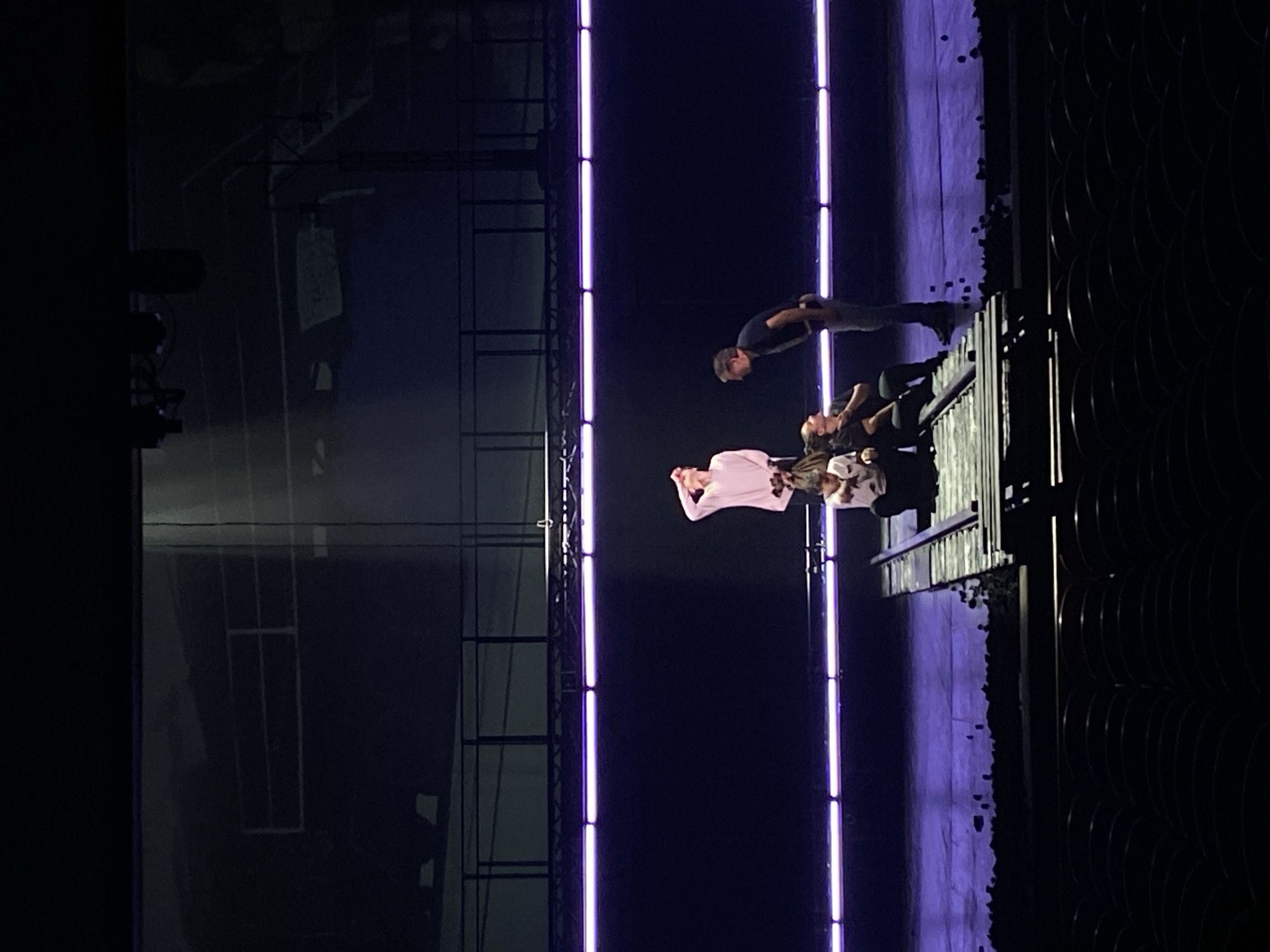 The image shows a stage performance with four people. Two vertical neon lights are positioned on the stage. The performers are standing on a raised platform with a black metal railing. The person on the left is wearing a light pink shirt, the next two individuals are wearing dark shirts with lighter details, and the person on the right is wearing a dark shirt and pants. The location is unknown.