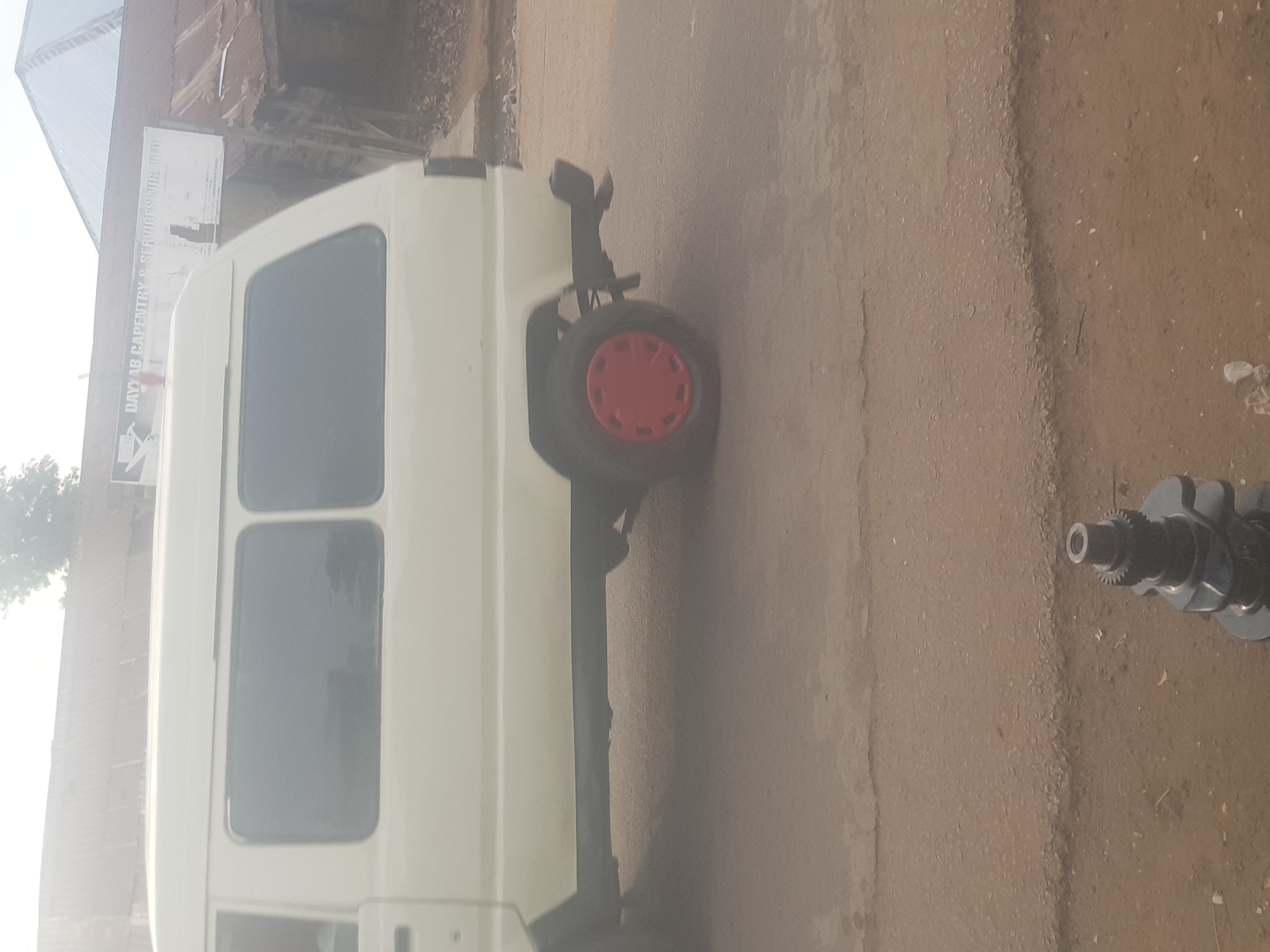 The image displays an outdoor scene in Kufena, Nigeria, featuring a white commercial-style van parked on a dusty, unpaved surface. The van, positioned on the left and central parts of the frame, shows its passenger side with multiple dark-tinted rectangular windows.
Towards the rear of the van, a single wheel with a black tire and a distinct red center cap is visible on the ground. This wheel appears to be mounted on a dark frame or stand, suggesting it might be a spare or part of a separate piece of equipment. Further to the right, resting on the dusty ground, is a dark metallic engine component resembling a crankshaft, identifiable by its lobed structure and a geared end.
In the background, behind the van, stands a building with a corrugated metal roof and what appears to be brick or mud walls. A white rectangular sign is mounted on this building, bearing partially legible text: "DAYLAB CARPENTRY & SERVICES NIG. LTD." The scene is brightly lit, indicating it is daytime. No people are visible, and no specific activity is occurring; the van is stationary, and the various mechanical parts are simply lying on the earthy ground.