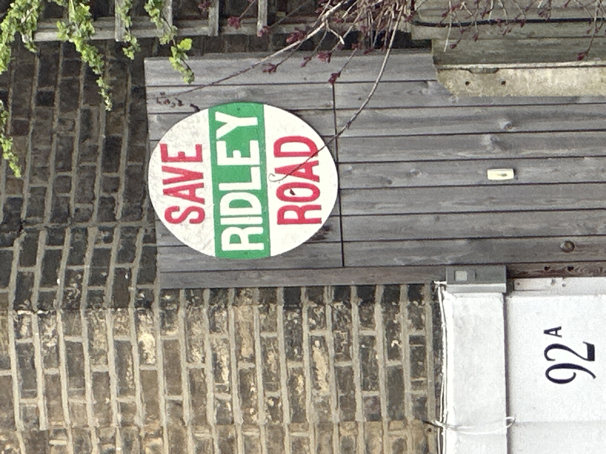 A daytime shot presents a brick facade with a "Save Ridley Road" sign affixed to wooden planking. The sign is circular with the words "SAVE" and "ROAD" in red, separated by the name "RIDLEY" in green. The address "92A" is printed on a white surface. This scene is located in Ridley Road, London, United Kingdom.