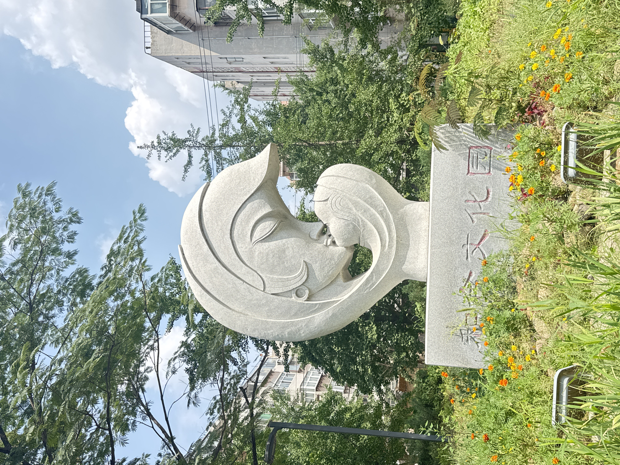 This is a medium shot photograph of a stone sculpture of a mother and child in the city of Zhongtun, China. The sculpture is white and abstract, depicting a mother kissing her child. It is situated in a park or garden area with lush green foliage and small orange and yellow flowers. Behind the sculpture, there are several buildings, some of which appear to be apartment complexes, and trees with dense green leaves. The sky is blue with some white clouds. The overall scene suggests a peaceful and family-oriented atmosphere.